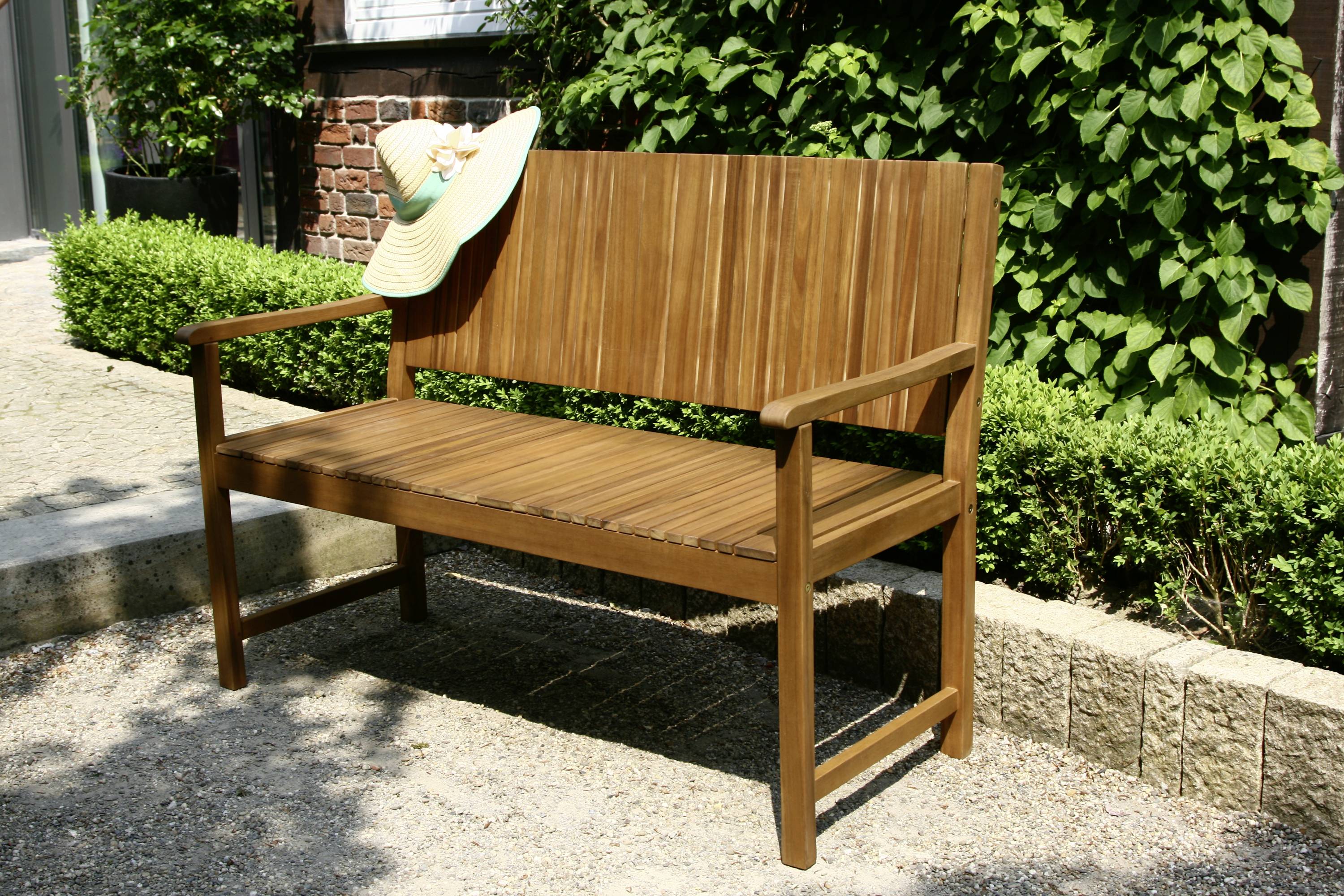 Holzbank im sonnigen Garten, auf der ein Sonnenhut liegt. Im Hintergrund sind grüne Sträucher und eine Ziegelmauer zu sehen.