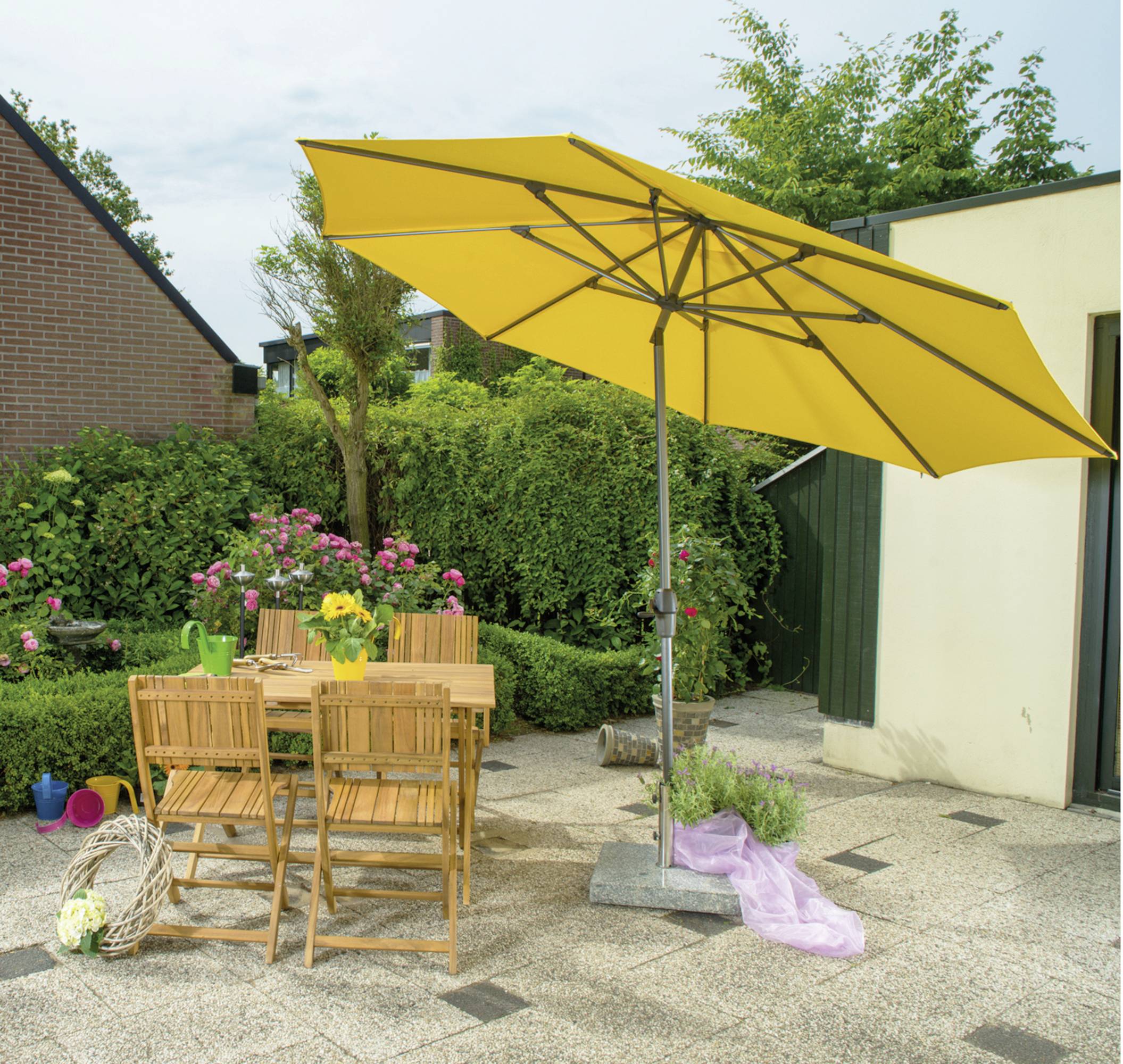 Gelber Sonnenschirm über einem Holztisch mit vier Stühlen auf einer Terrasse. Im Hintergrund grüner Garten mit Blumenbeeten.