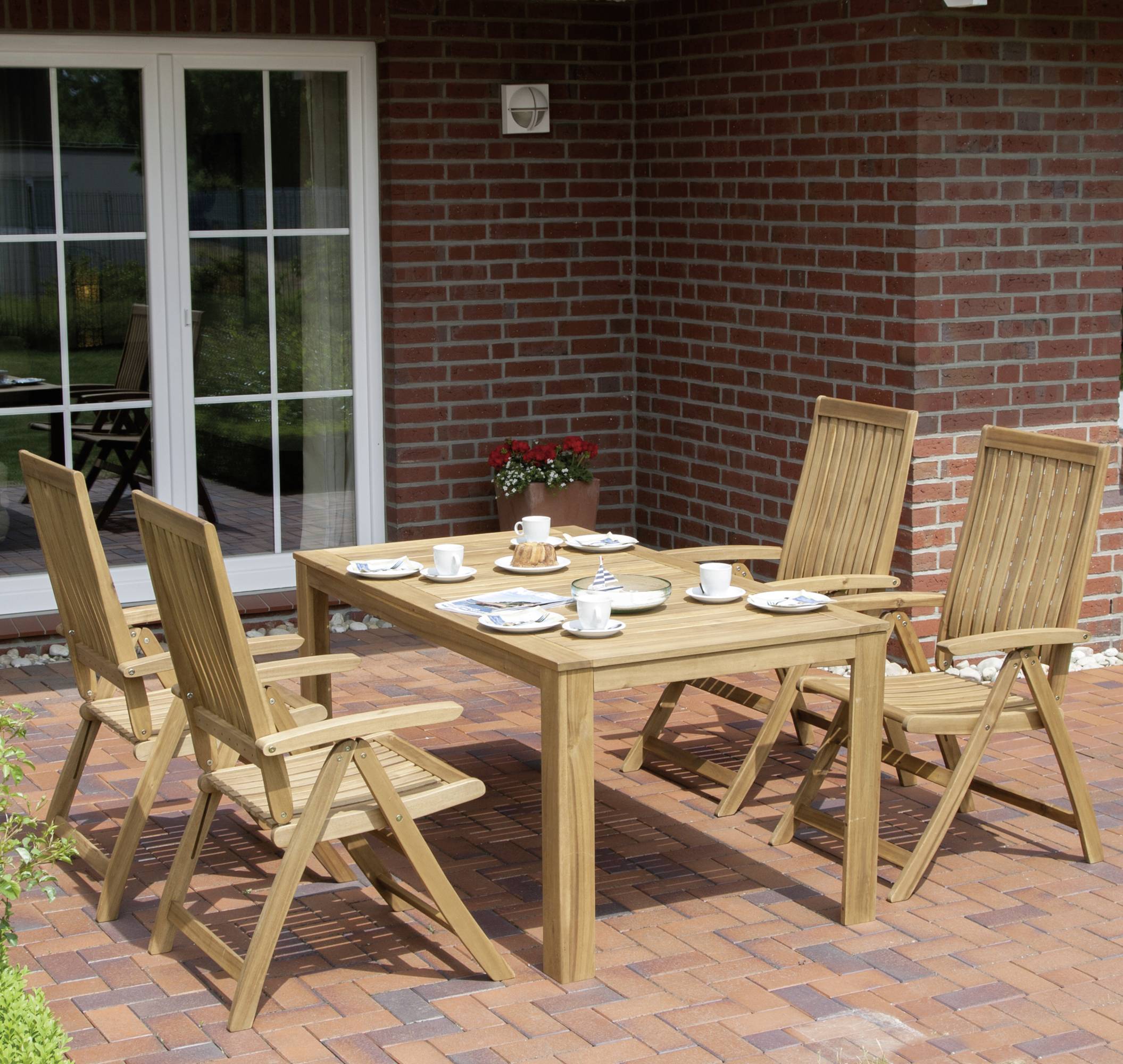 Gartentisch mit vier Holzbänken, gedeckt mit Geschirr und Tassen, auf einer Ziegelterrasse vor einer Backsteinfassade mit Glastür.