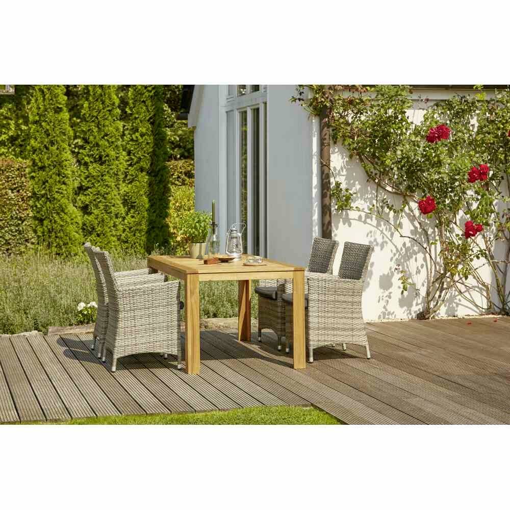 Ein Holztisch mit zwei geflochtenen Stühlen steht auf einer Terrasse neben einem weißen Haus und blühenden Rosensträuchern.