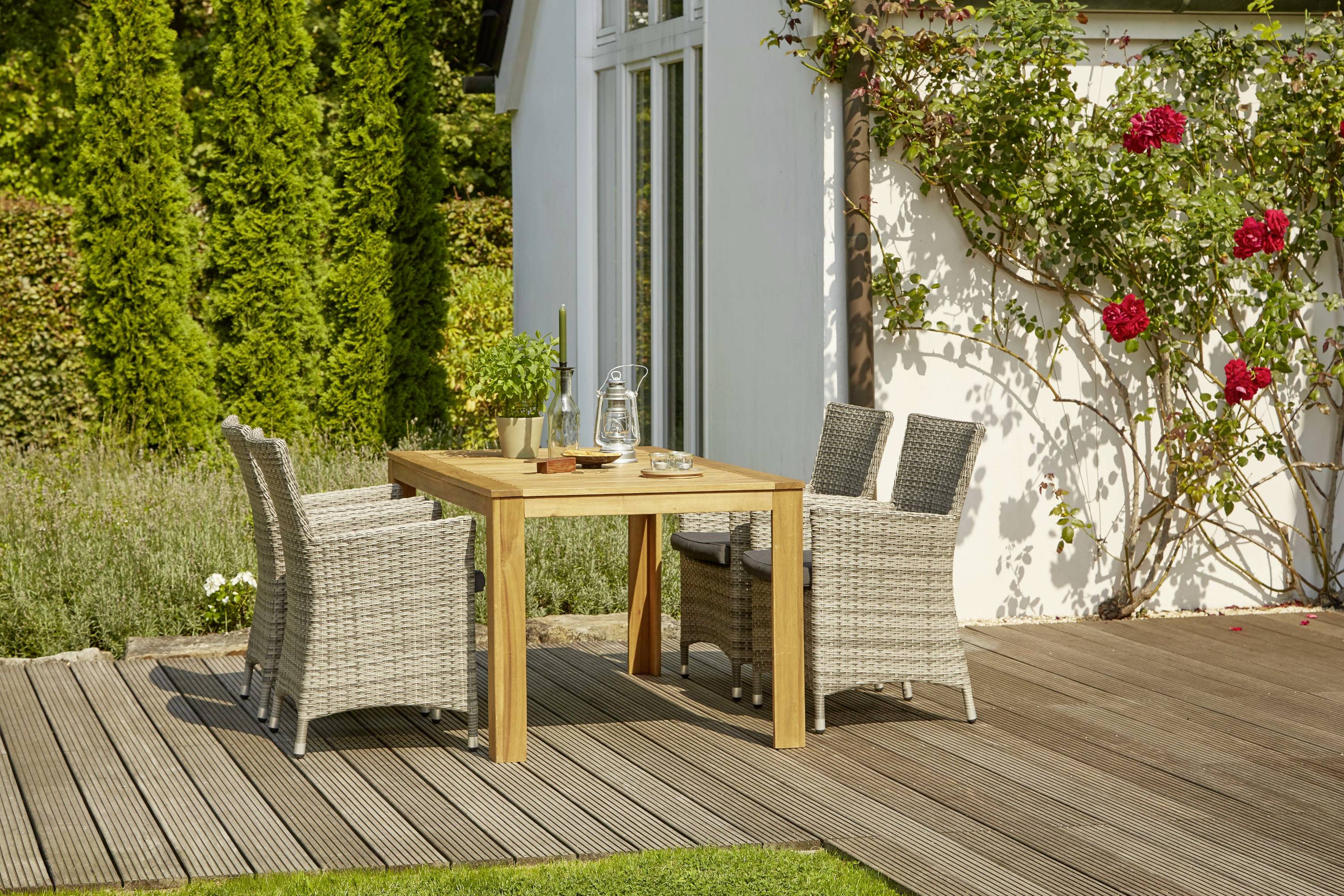 Ein Holztisch auf einer Terrasse, umgeben von vier geflochtenen Stühlen, flankiert von einem grünen Garten und blühenden roten Rosen.