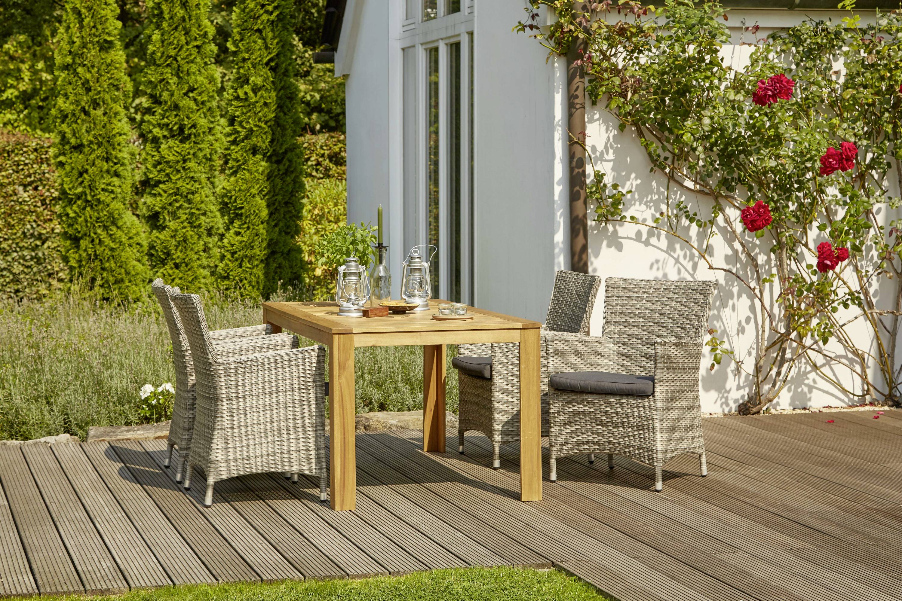 Ein Holztisch mit zwei geflochtenen Stühlen steht auf einer Terrasse. Im Hintergrund sind ein Gartenhaus, grüne Büsche und rote Rosen zu sehen.