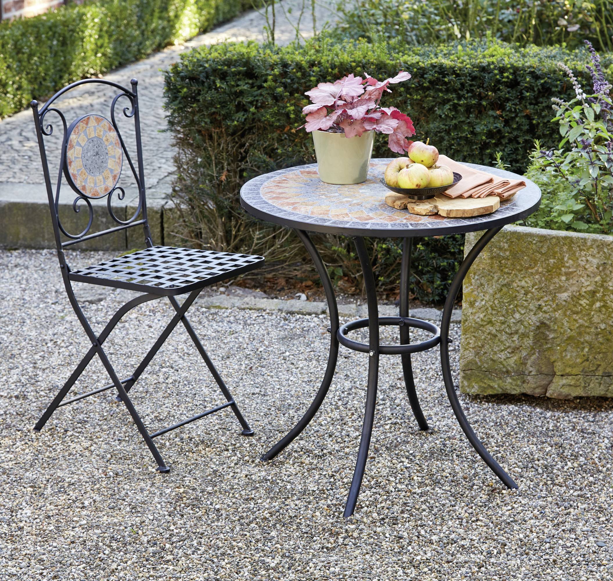 Schwarzer Metalltisch mit Mosaikplatte; darauf dekorative Pflanzen und Äpfel. Daneben passender Stuhl, auf einem Kiesweg im Garten.