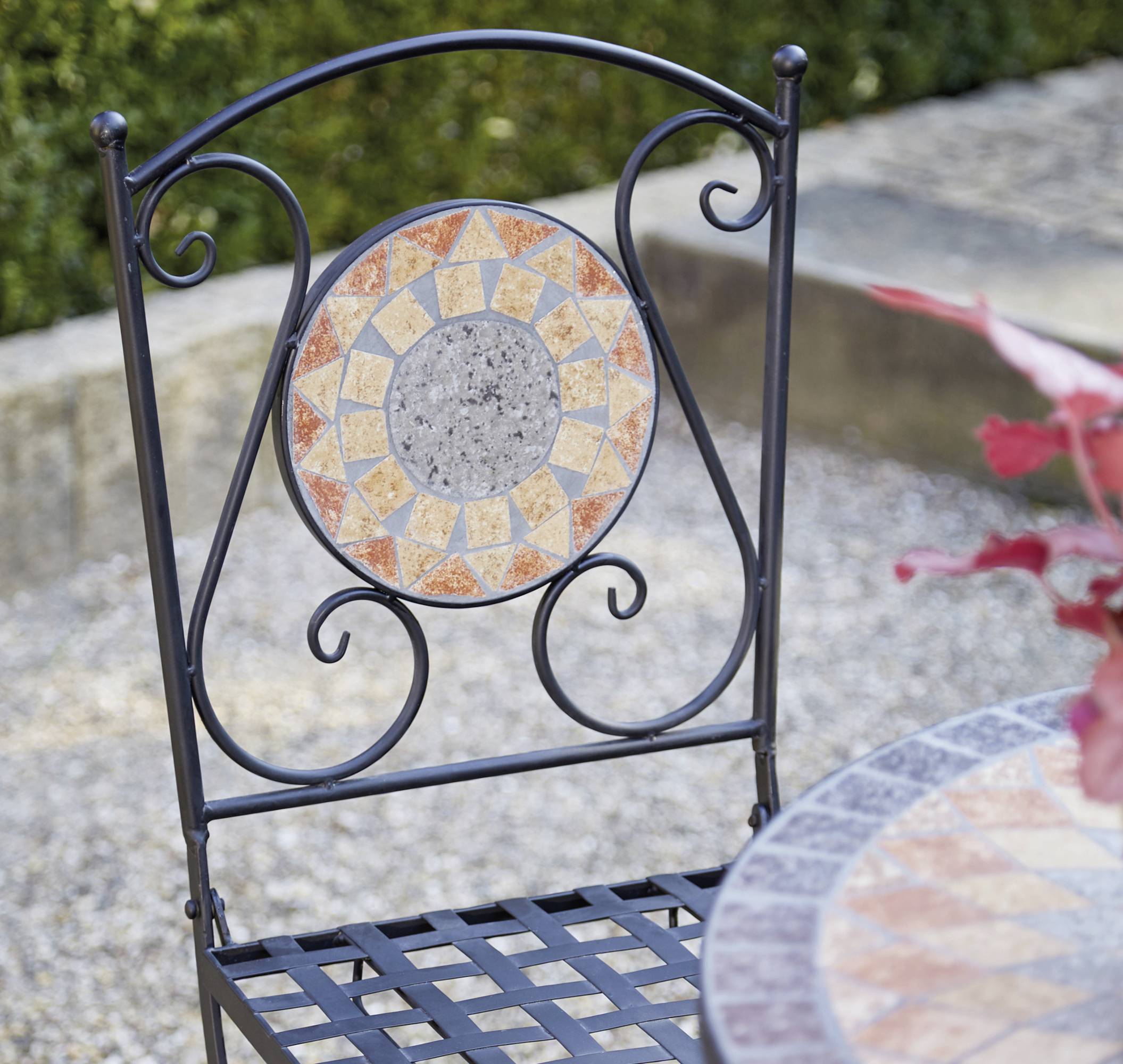 Schwarzer Metallstuhl mit dekorativem Mosaikmuster in einem Garten auf Kiesboden, neben einem Tisch mit ähnlichem Design.