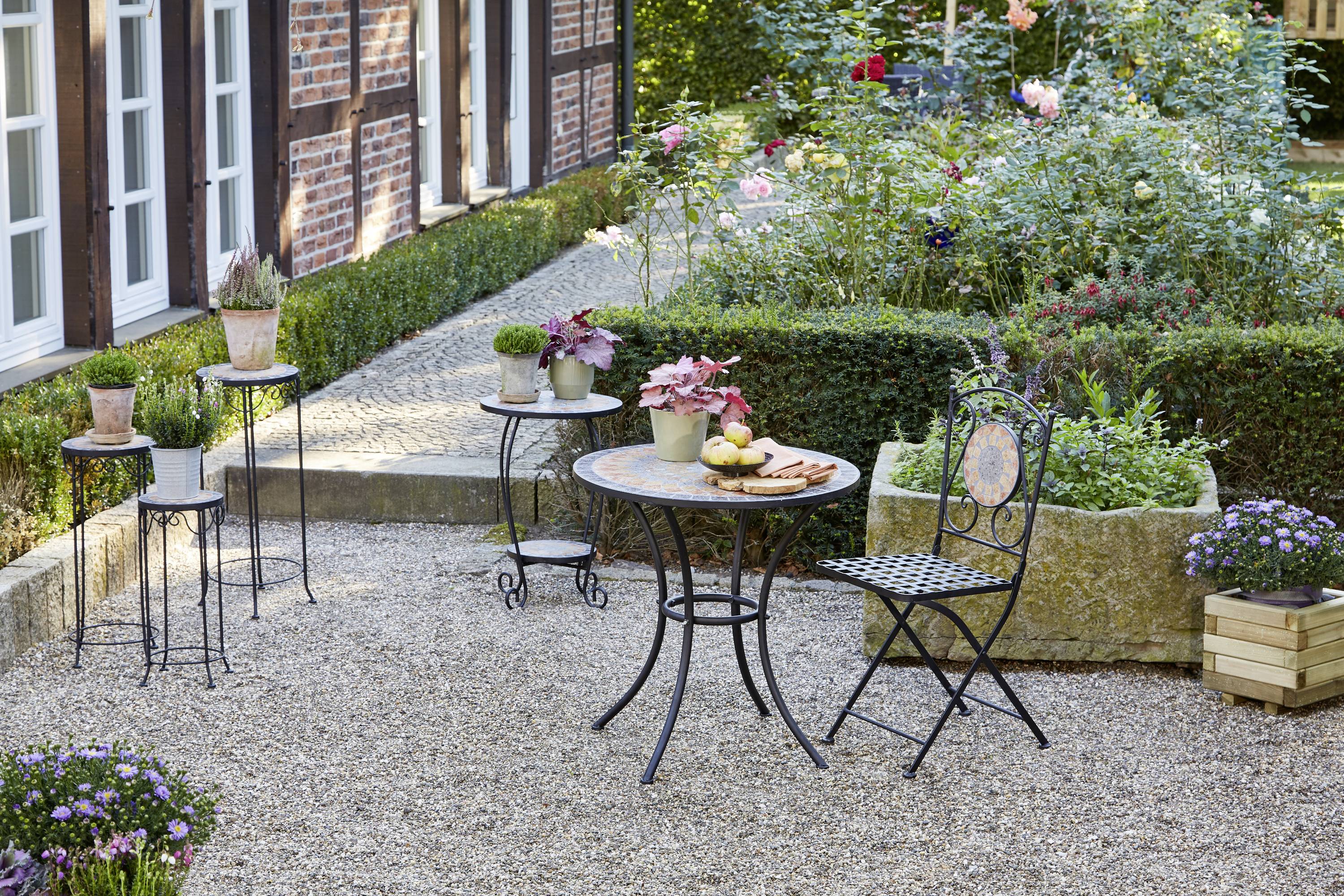 Ein kleiner Garten mit Kiesweg, Tisch und Stühlen im Vordergrund, umgeben von blühenden Blumen und Hecken. Rustikales Ambiente.