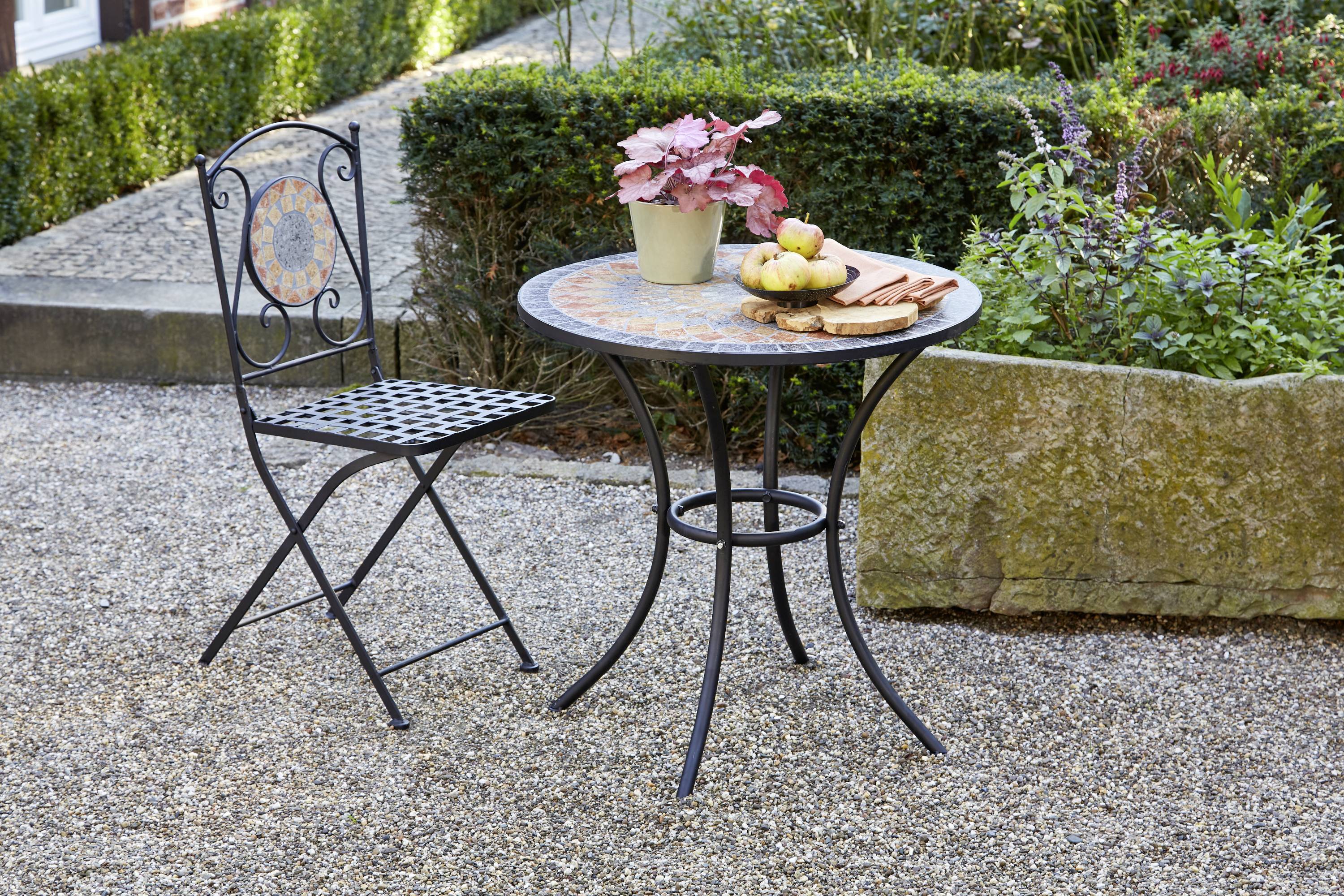 Ein Gartentisch mit Mosaikmuster steht auf Kies. Auf dem Tisch sind ein Blumentopf mit roten Blättern und einige Äpfel platziert.