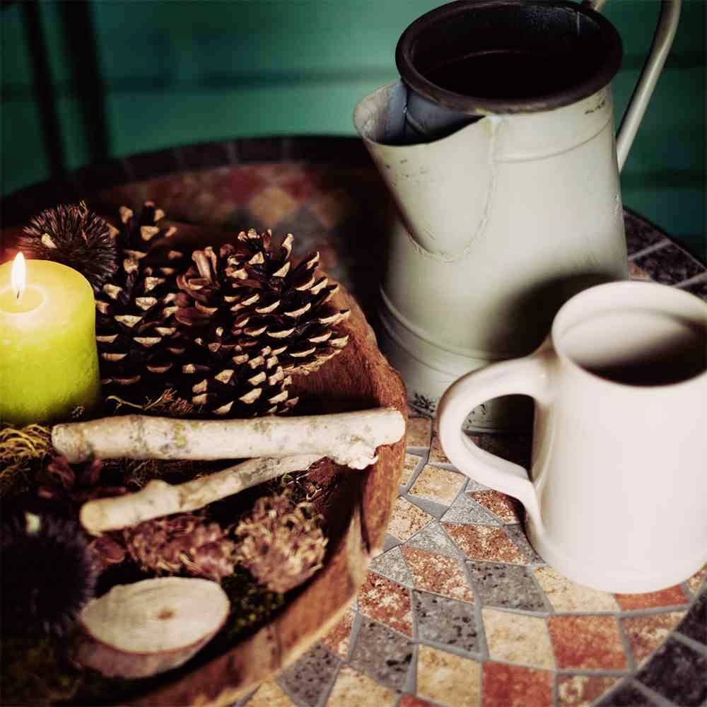 Ein dekorativer Tisch mit einer brennenden Kerze, Kiefernzapfen, Zweigen und einem abgenutzten Krug sowie einer Tasse.