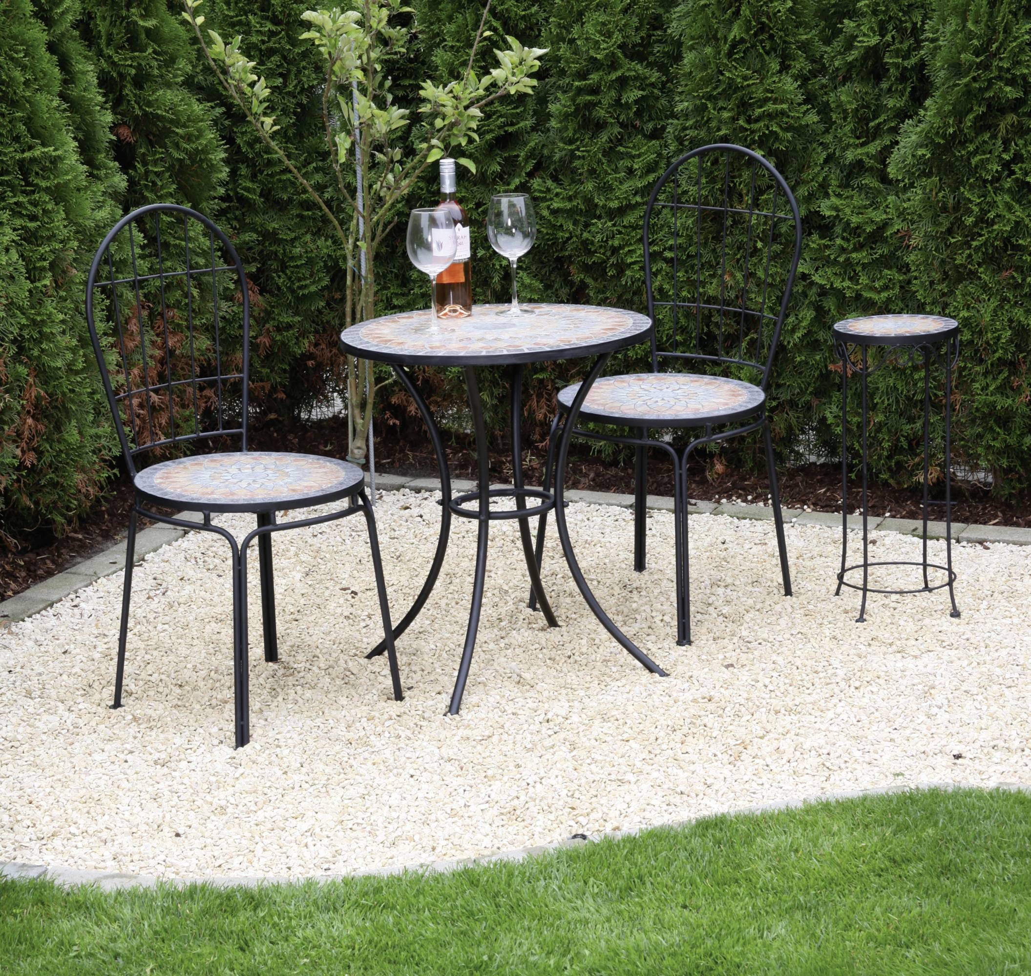 Garten mit zwei Stühlen und kleinem Tisch aus Metall auf Kiesboden, Flasche Roséwein und zwei Gläser darauf, umgeben von Hecken.