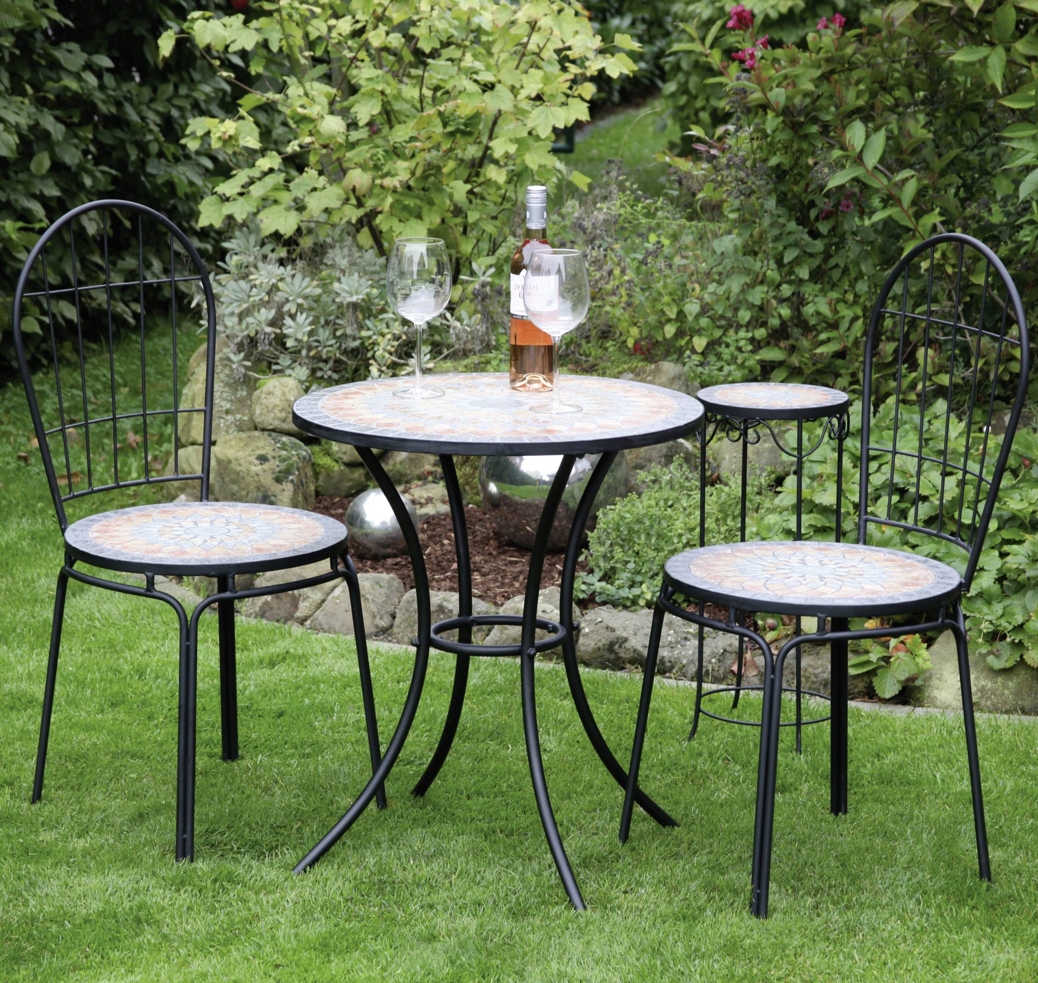 Runder Tisch mit zwei Stühlen auf Rasenfläche in einem Garten, darauf eine Flasche Wein und zwei Gläser. Im Hintergrund blühende Pflanzen.