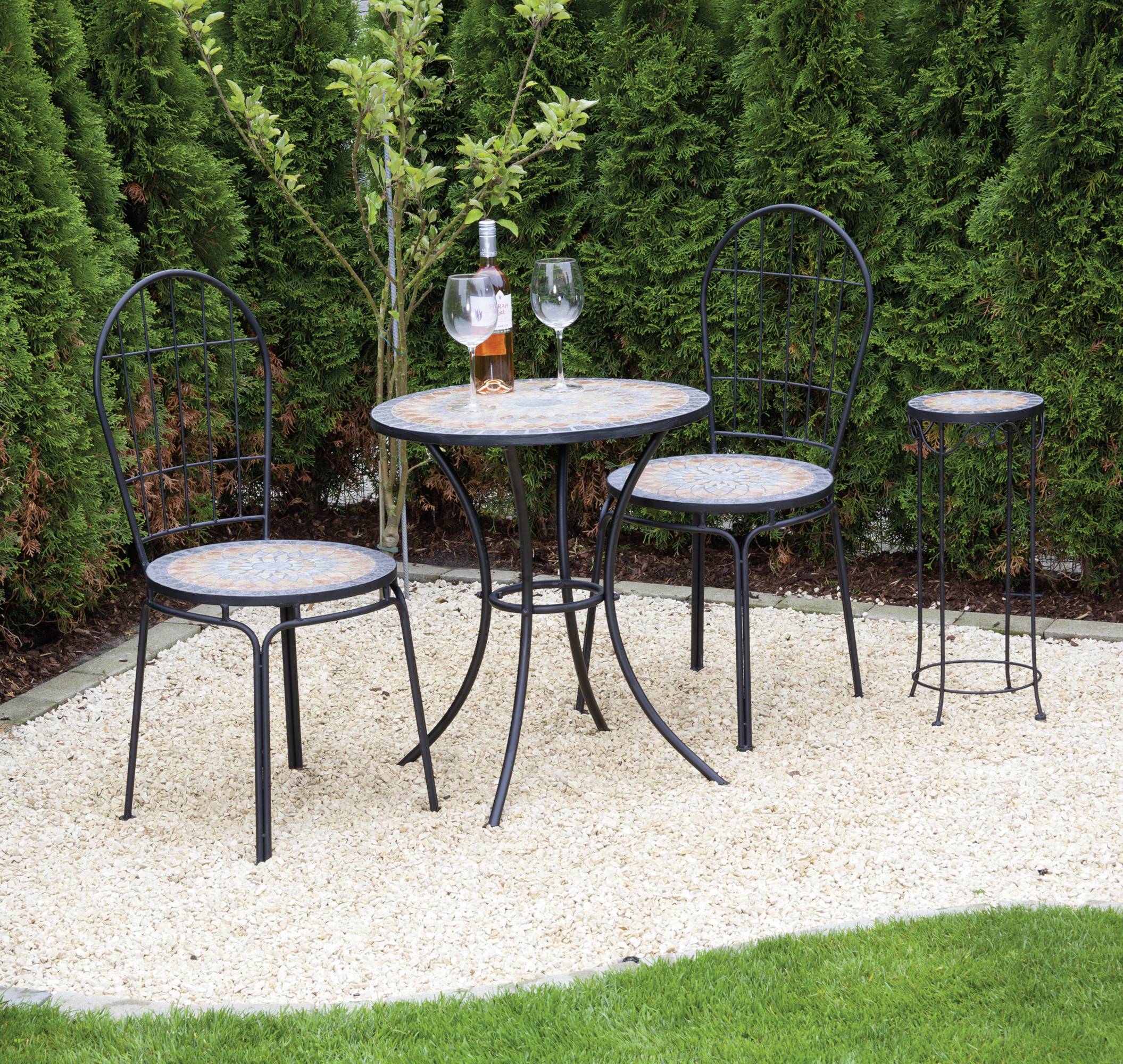 Ein kleiner Gartentisch mit zwei Stühlen und einem Hocker, auf dem Tisch stehen eine geöffnete Weinflasche und zwei Gläser, umgeben von grünen Pflanzen im Freien.