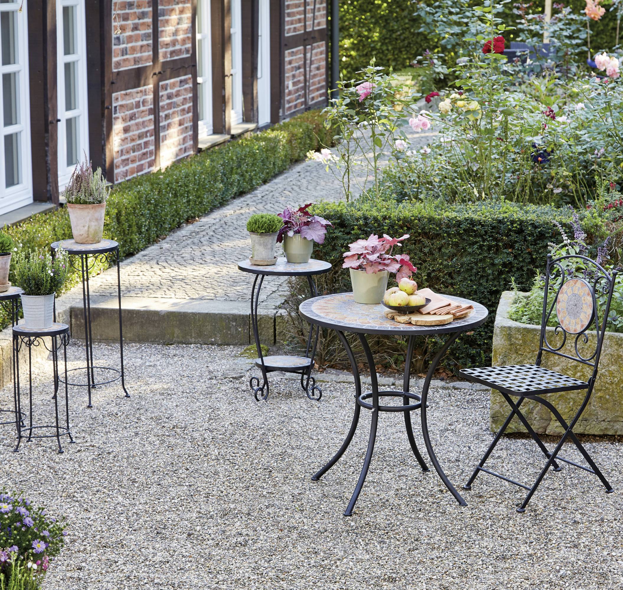 Ein gemütlicher Gartenbereich mit einem runden Tisch, dekorativen Metallsesseln und Pflanzen. Pflastersteinweg und blühende Rosen im Hintergrund.