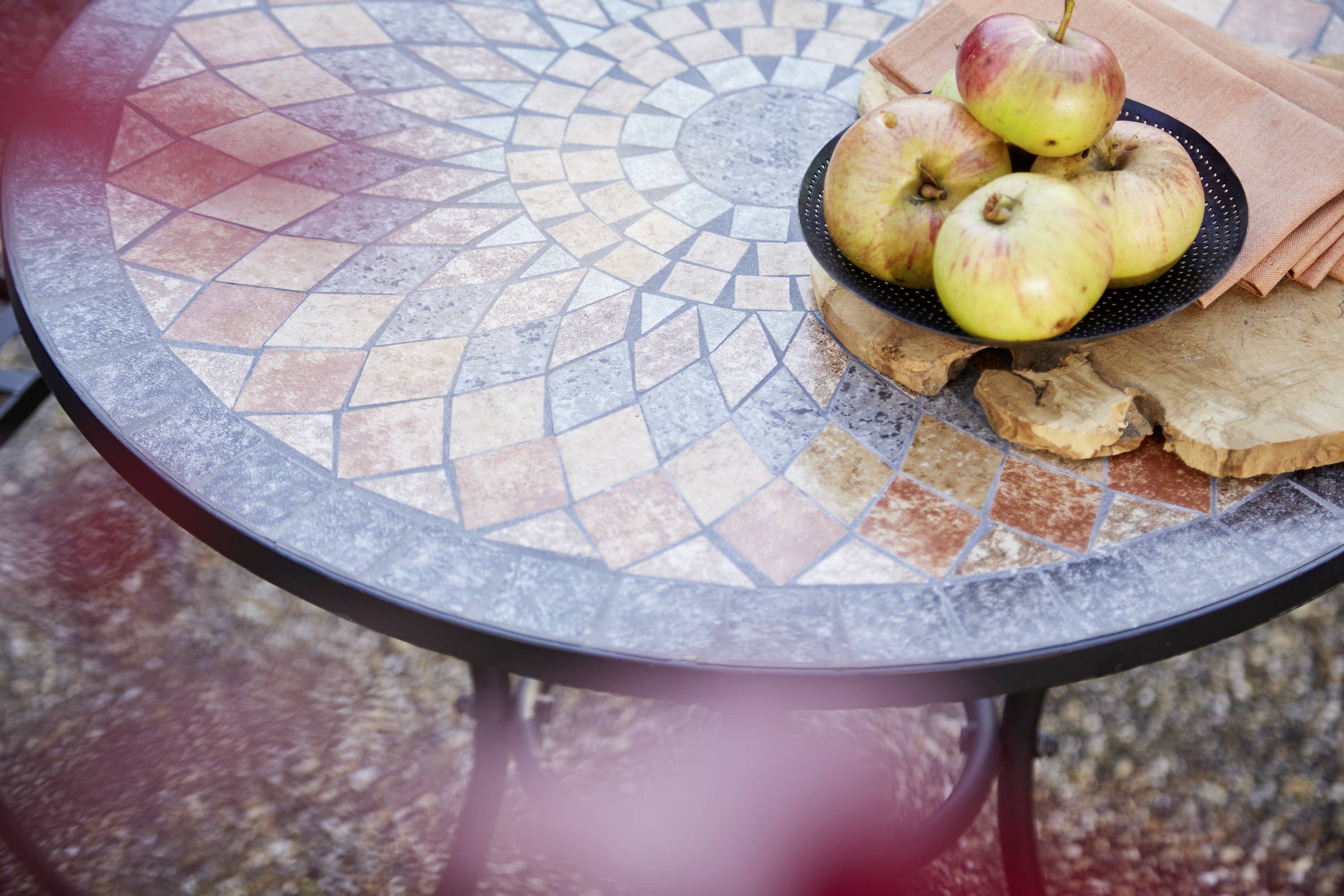 Runder, mosaikverzierter Tisch mit einem Teller voller Äpfel und einem Serviettenset, auf einem Holzbrett liegend.