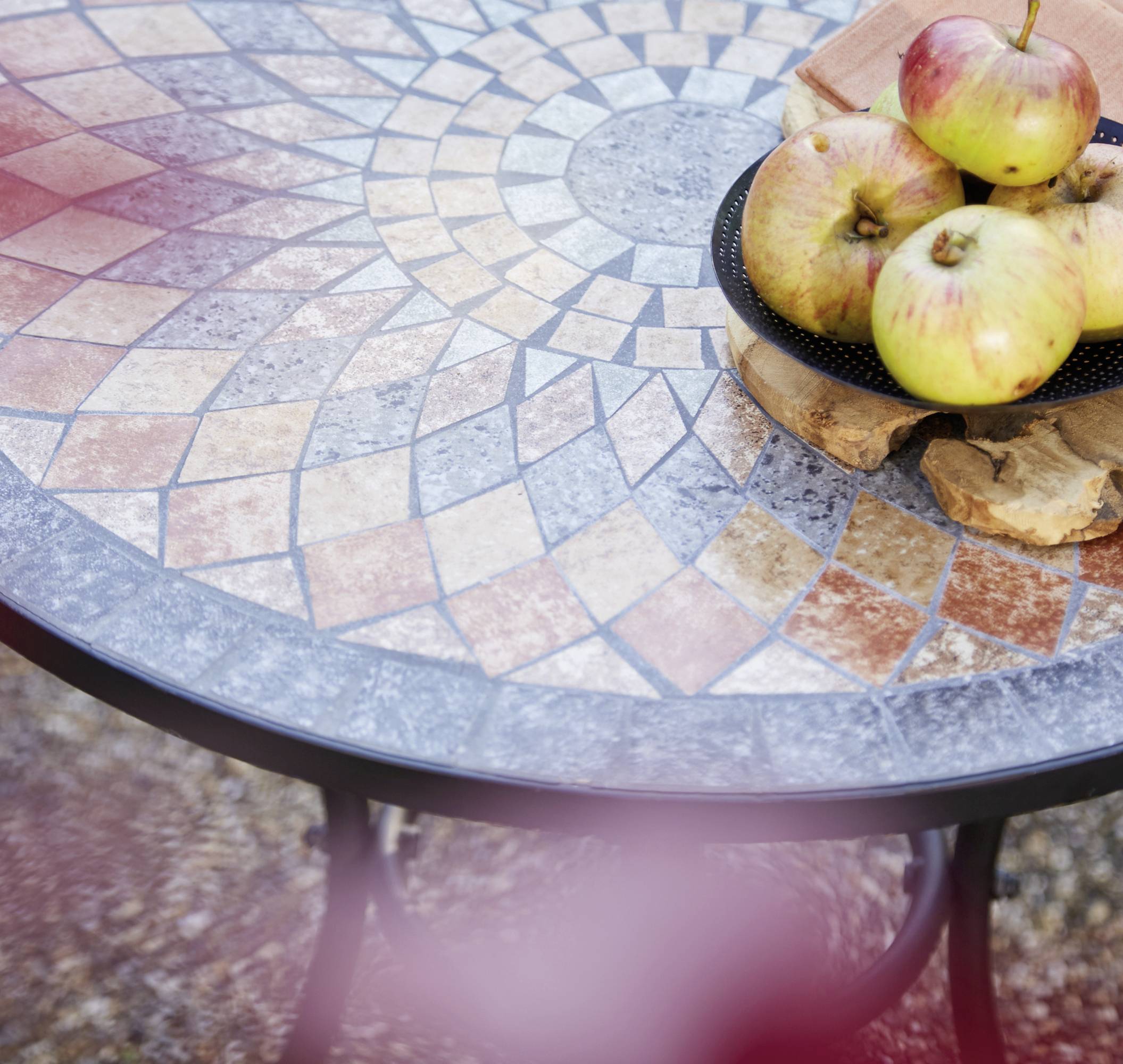 Runder Tisch mit Mosaikfliesen in verschiedenen Farben. Darauf liegt ein Holzbrett mit mehreren gelben Äpfeln. Im Hintergrund unscharf.