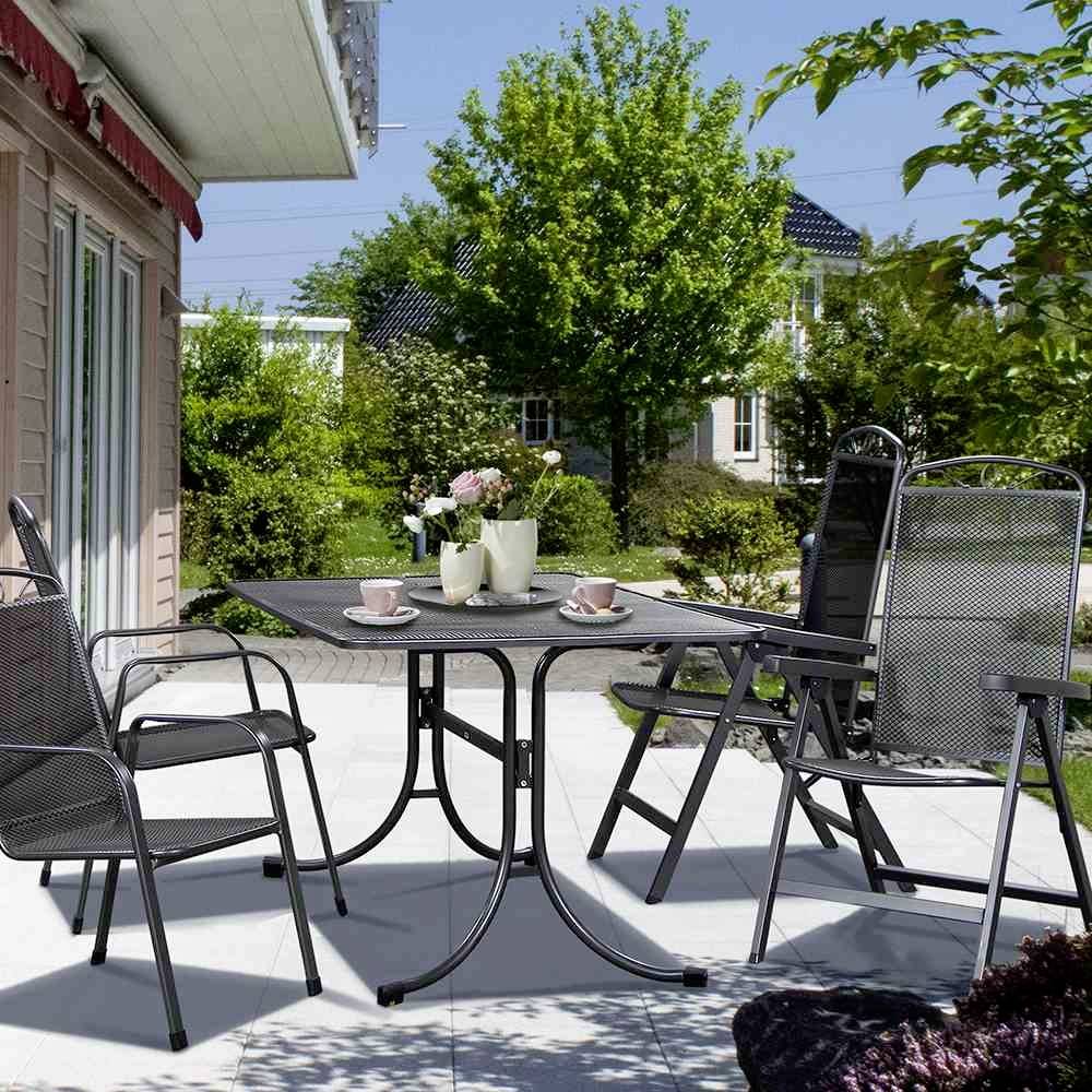 Ein Tisch mit vier Stühlen auf einer Terrasse, umgeben von grünem Garten. Auf dem Tisch stehen Blumen und Kaffeetassen.