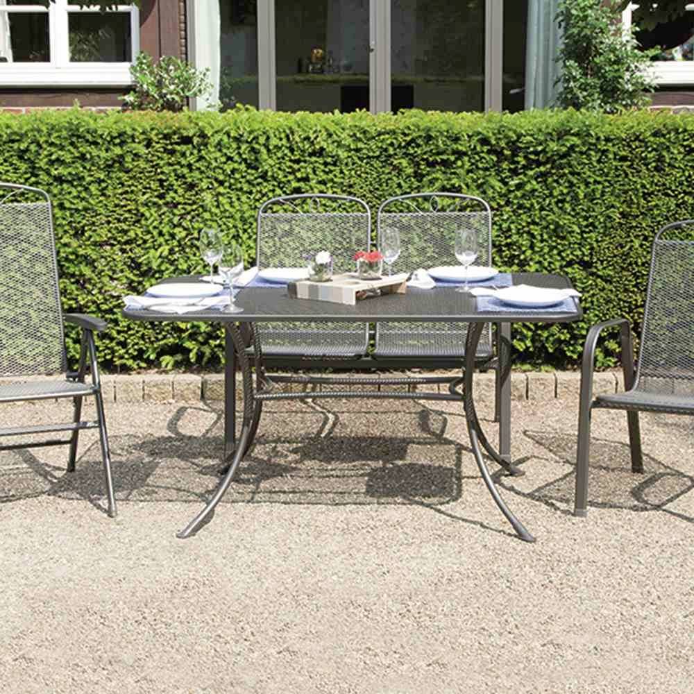 Gartenmöbel-Set auf einem Kiesweg vor einer Hecke. Metalltisch mit vier Stühlen, Gedeck und Blumendekoration in der Mitte.