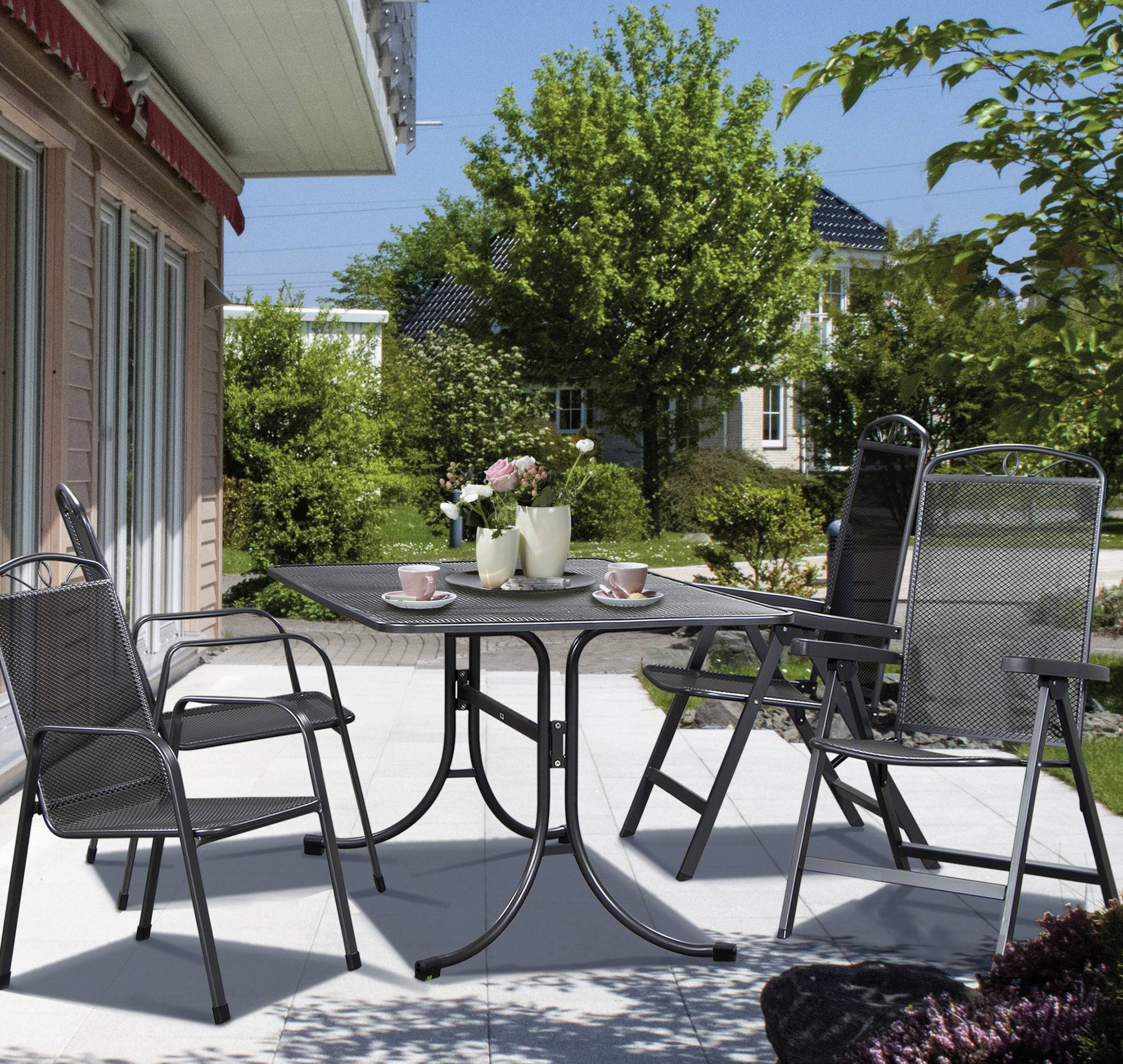 Ein Garten mit Tisch und vier Stühlen. Auf dem Tisch stehen Tassen und eine Vase mit Blumen. Im Hintergrund sind Bäume und Sträucher.