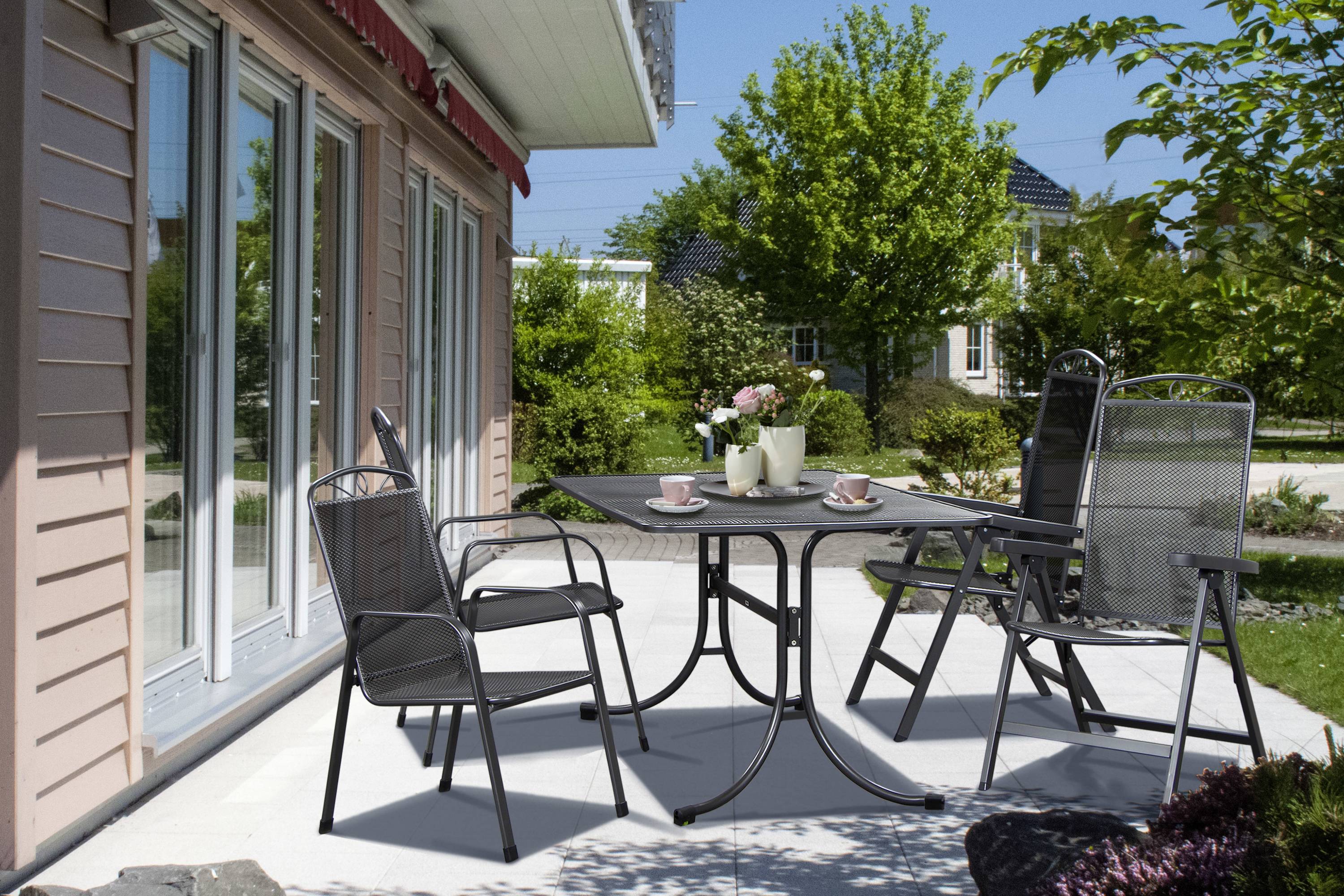 Ein Tisch mit vier leeren Stühlen auf einer Terrasse neben einem Haus. Auf dem Tisch stehen eine Kaffeekanne, Tassen und Blumen.