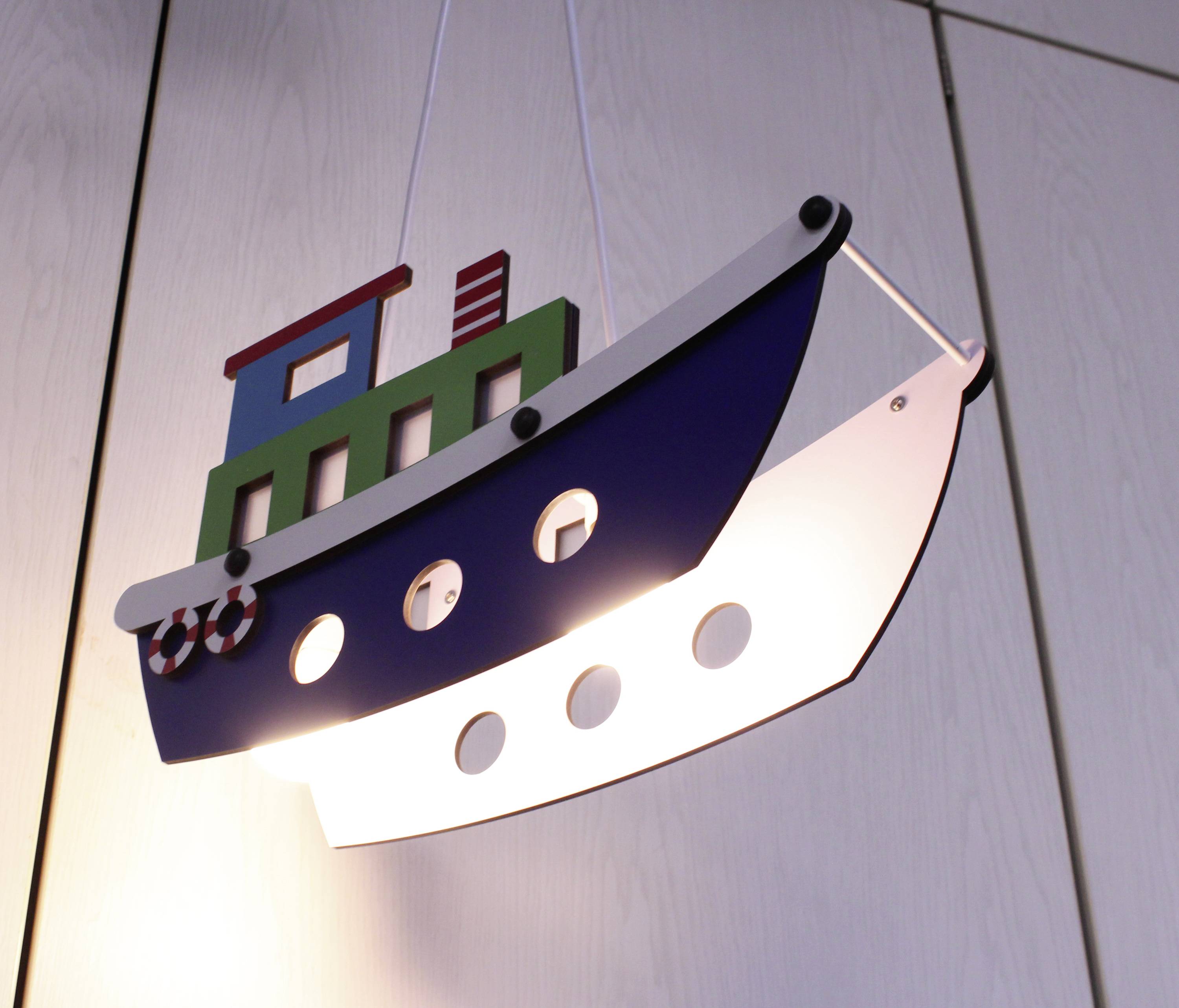 Eine Hängelampe in Form eines Schiffes mit Fenstern leuchtet. Sie hat bunte Details und hängt vor einer hellen Holzwand.