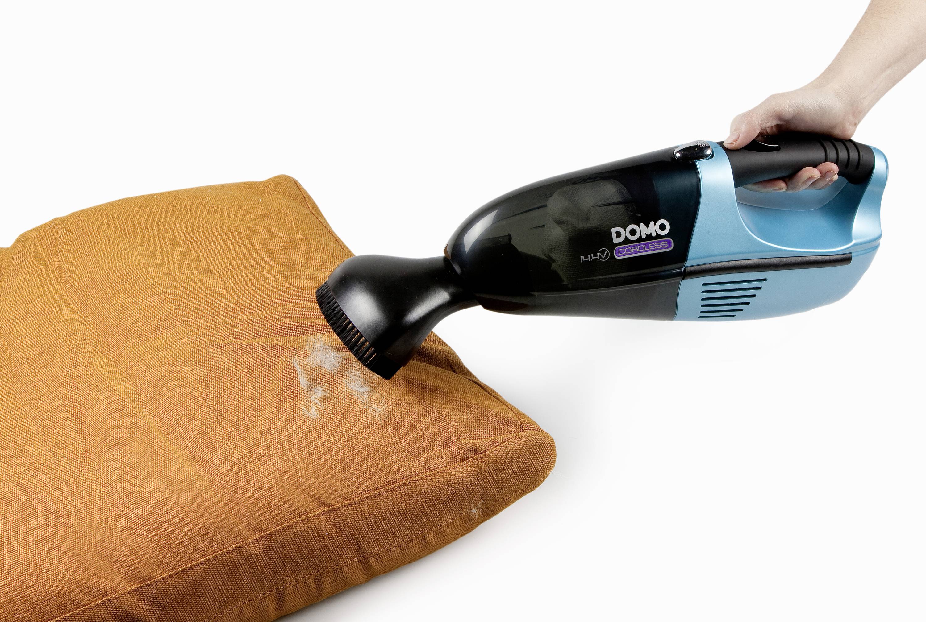 Eine Person hält einen blauen Handstaubsauger, der Staub von einem orangefarbenen Kissen saugt.