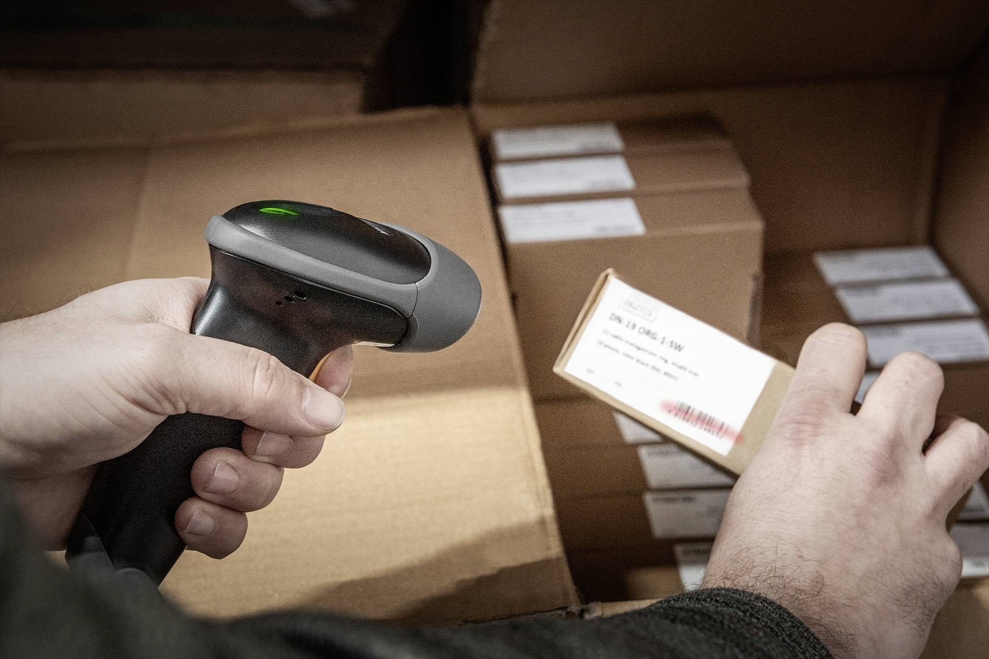 Eine Person scannt einen Barcode auf einer kleinen Schachtel mit einem Handscanner, umgeben von gestapelten Kartons im Lager.