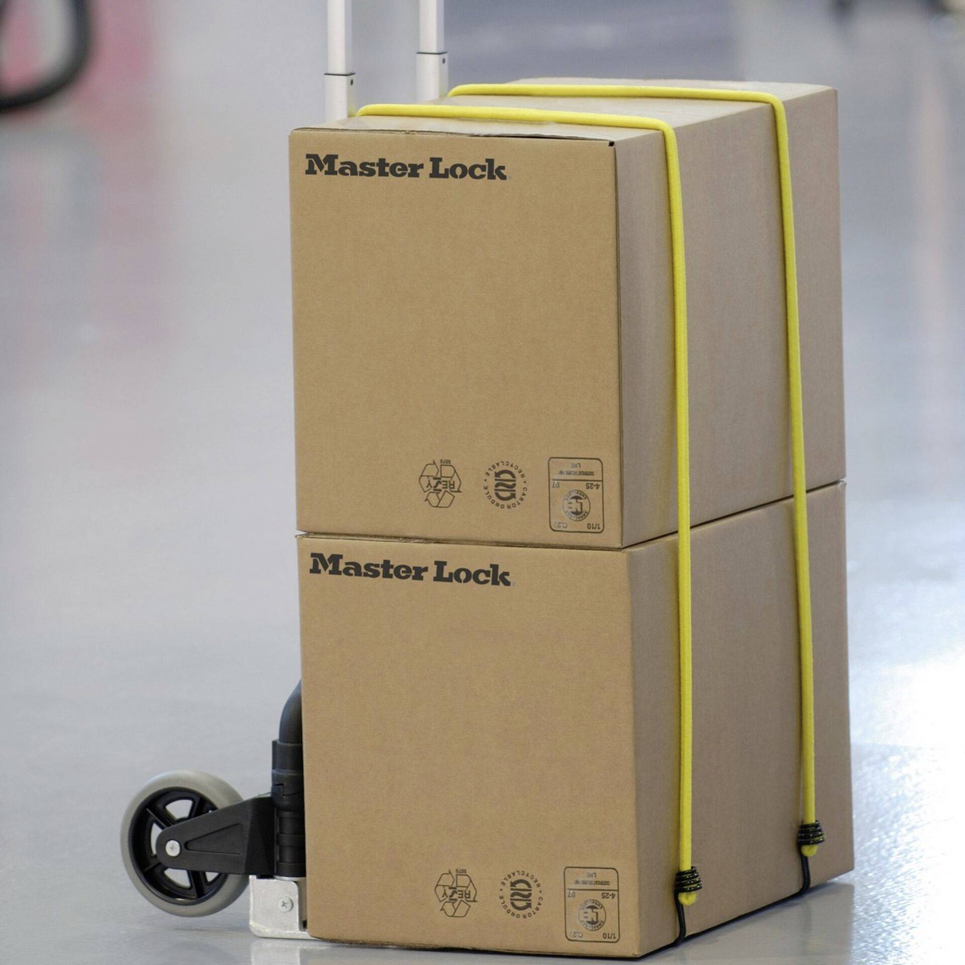 Ein Transportwagen trägt zwei gestapelte Kartons der Marke 'Master Lock', gesichert mit einem gelben Band und bereit für den Versand.