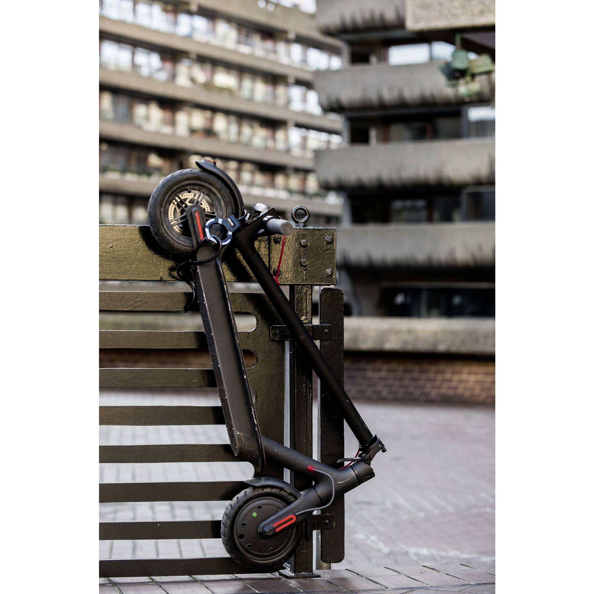 Ein zusammengeklappter E-Scooter lehnt gegen eine Parkbank in einer städtischen Umgebung. Im Hintergrund sind moderne Gebäude zu sehen.