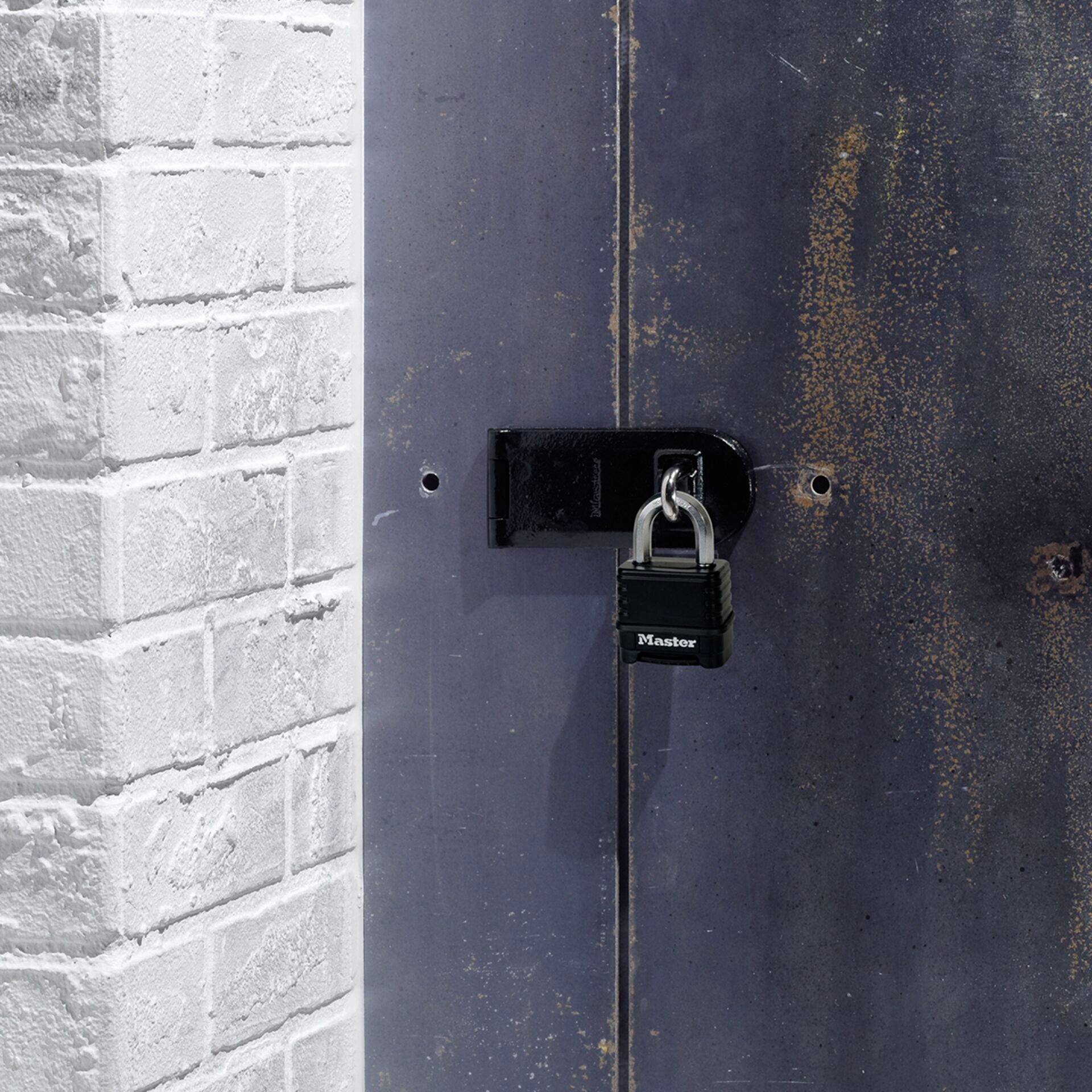 Metalltür mit Vorhängeschloss an einer weißen Backsteinmauer befestigt. Tür hat rostige Stellen, Schloss erscheint sicher.