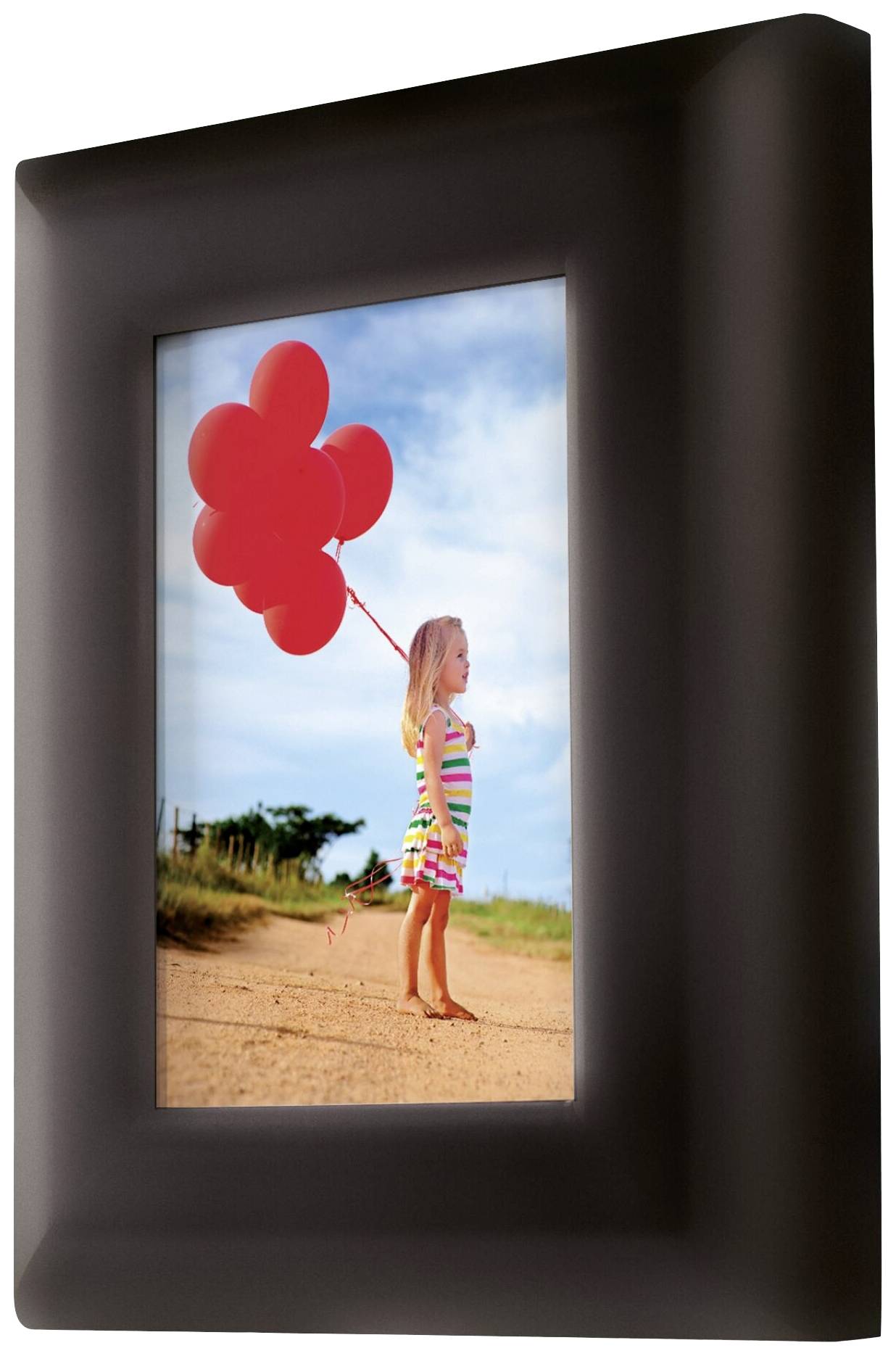 Ein kleines Mädchen in einem gestreiften Kleid hält rote Luftballons. Sie steht auf einem sandigen Weg unter blauem Himmel.