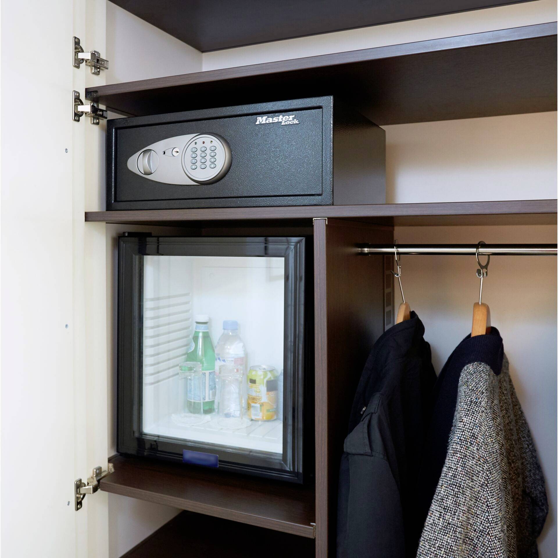 Ein geöffneter Kleiderschrank mit einem Safe oben, unter dem sich ein Mini-Kühlschrank mit Wasserflaschen und Getränken befindet. Rechts hängen Jacken.