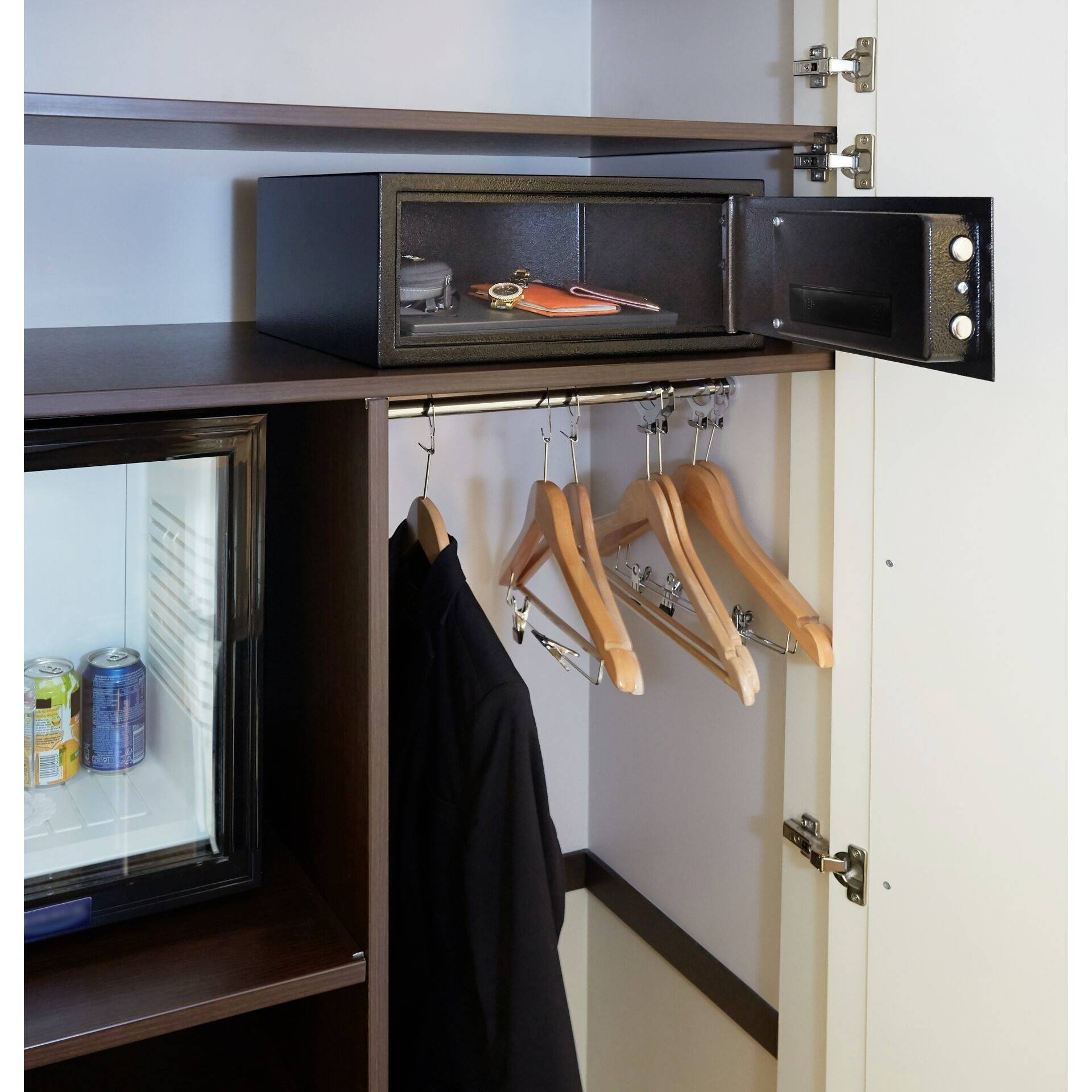 'Ein geöffneter Safe in einem Schrank, gefüllt mit persönlichen Gegenständen. Daneben Kleiderbügel mit Kleidung und ein Kühlschrank mit Getränkedosen.'