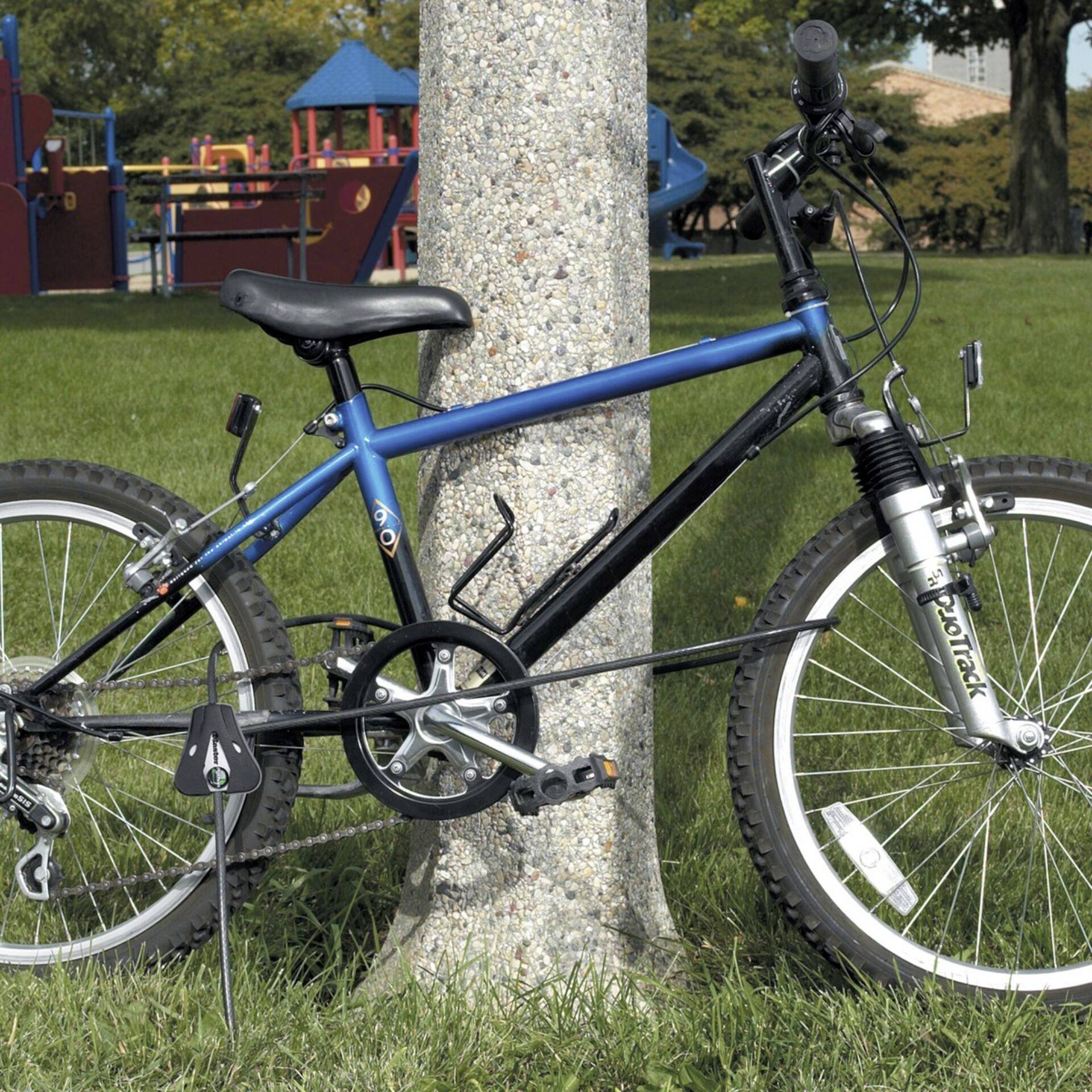 Ein blaues Fahrrad ist mit einem Schloss an einem Pfosten angekettet. Im Hintergrund befindet sich ein Kinderspielplatz auf einer grünen Wiese.