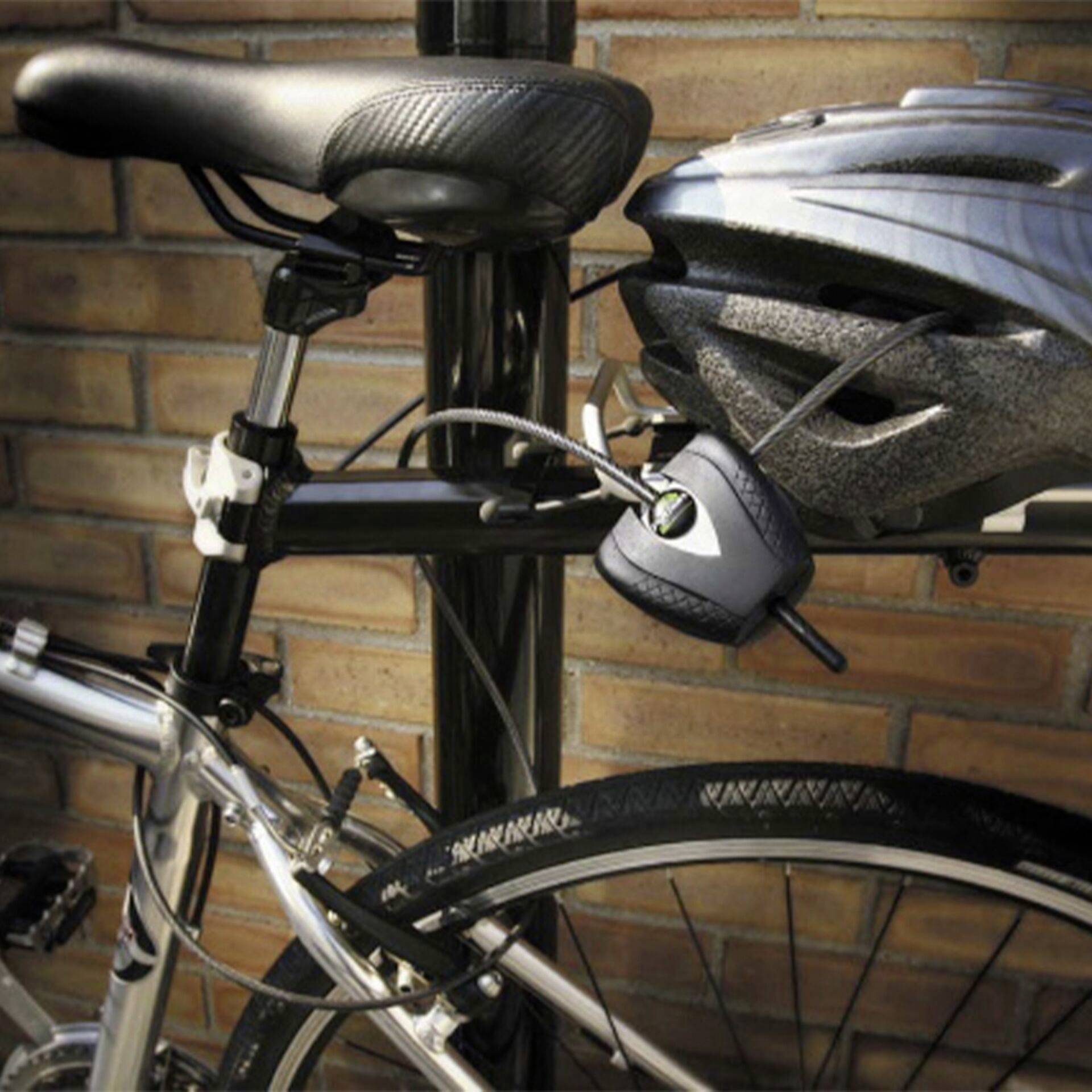 Ein Fahrrad mit Helm und Sicherheitskabel ist an einen Metallpfosten vor einer Ziegelwand angeschlossen.
