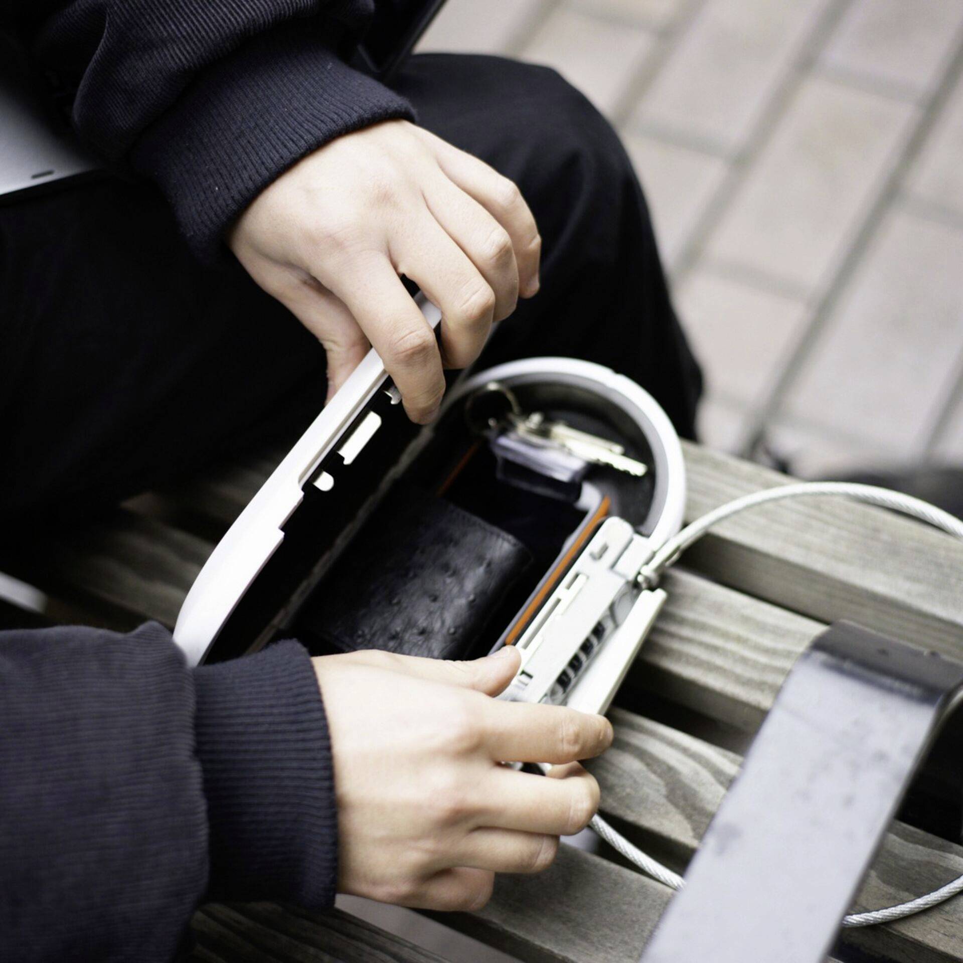 Eine Person öffnet eine weiße Schutzhülle auf einer Bank, um ein Smartphone aufzuladen. Ein Ladekabel ist angeschlossen.