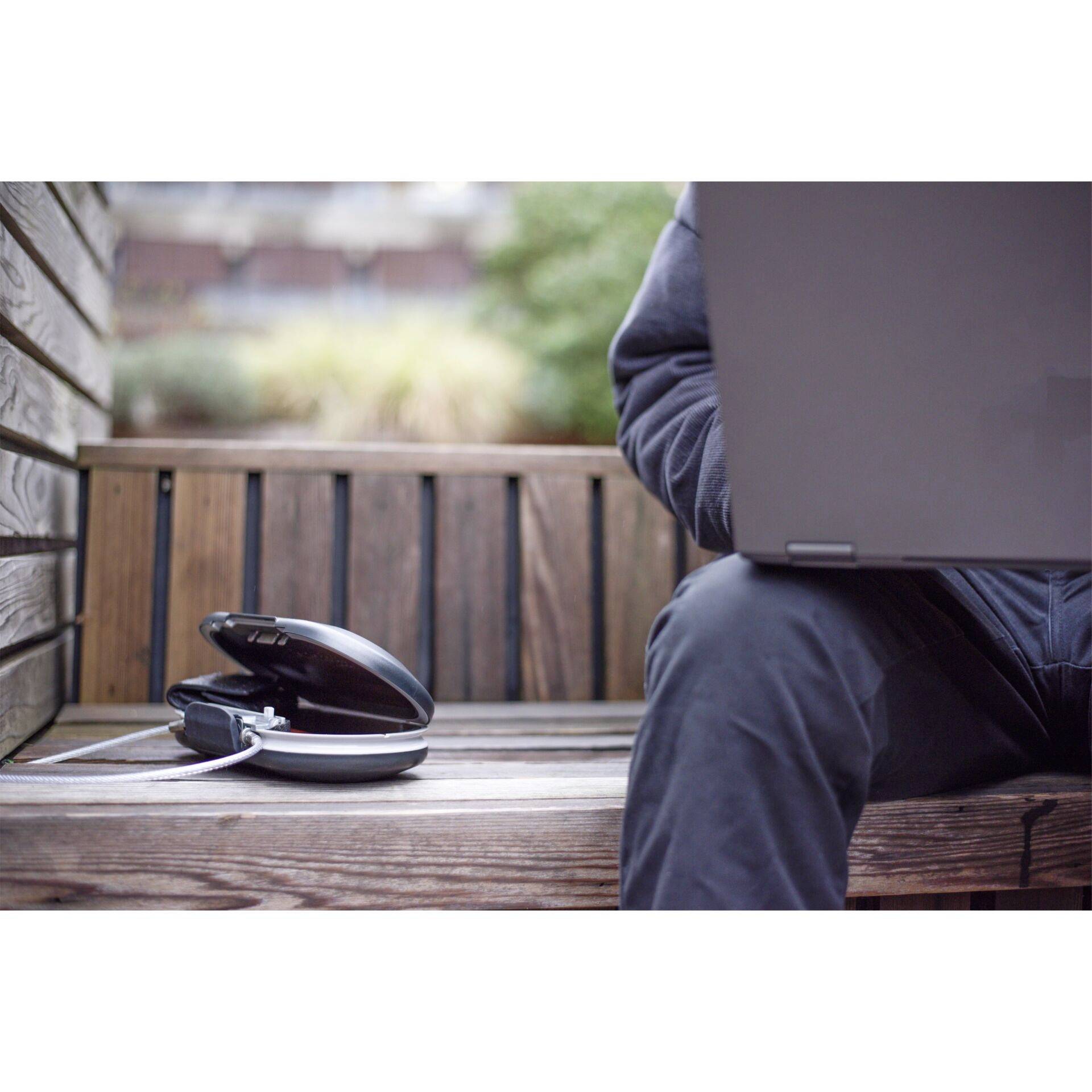 Eine Person sitzt auf einer Holzbank mit einem Laptop auf dem Schoß. Neben ihr liegen Kopfhörer. Die Umgebung ist unscharf.