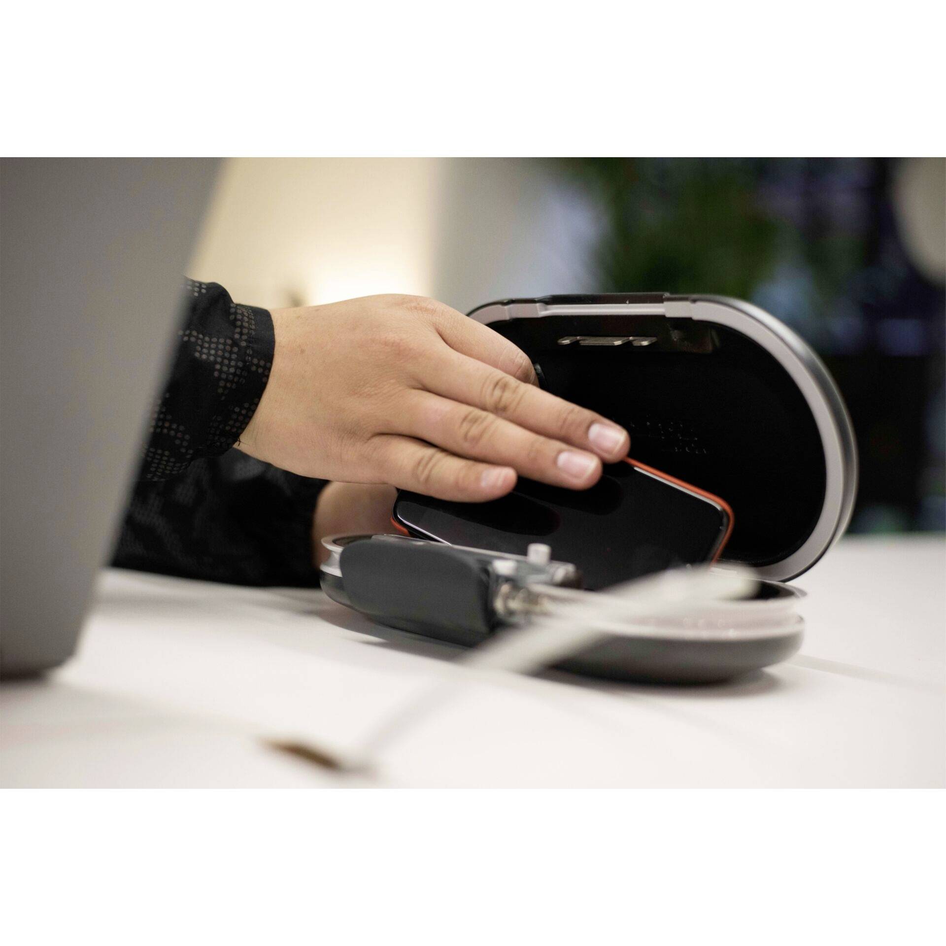 Eine Hand legt ein Smartphone in eine kabellose Ladestation auf einem Tisch. Im Hintergrund verschwommenes Licht und ein Laptop sichtbar.