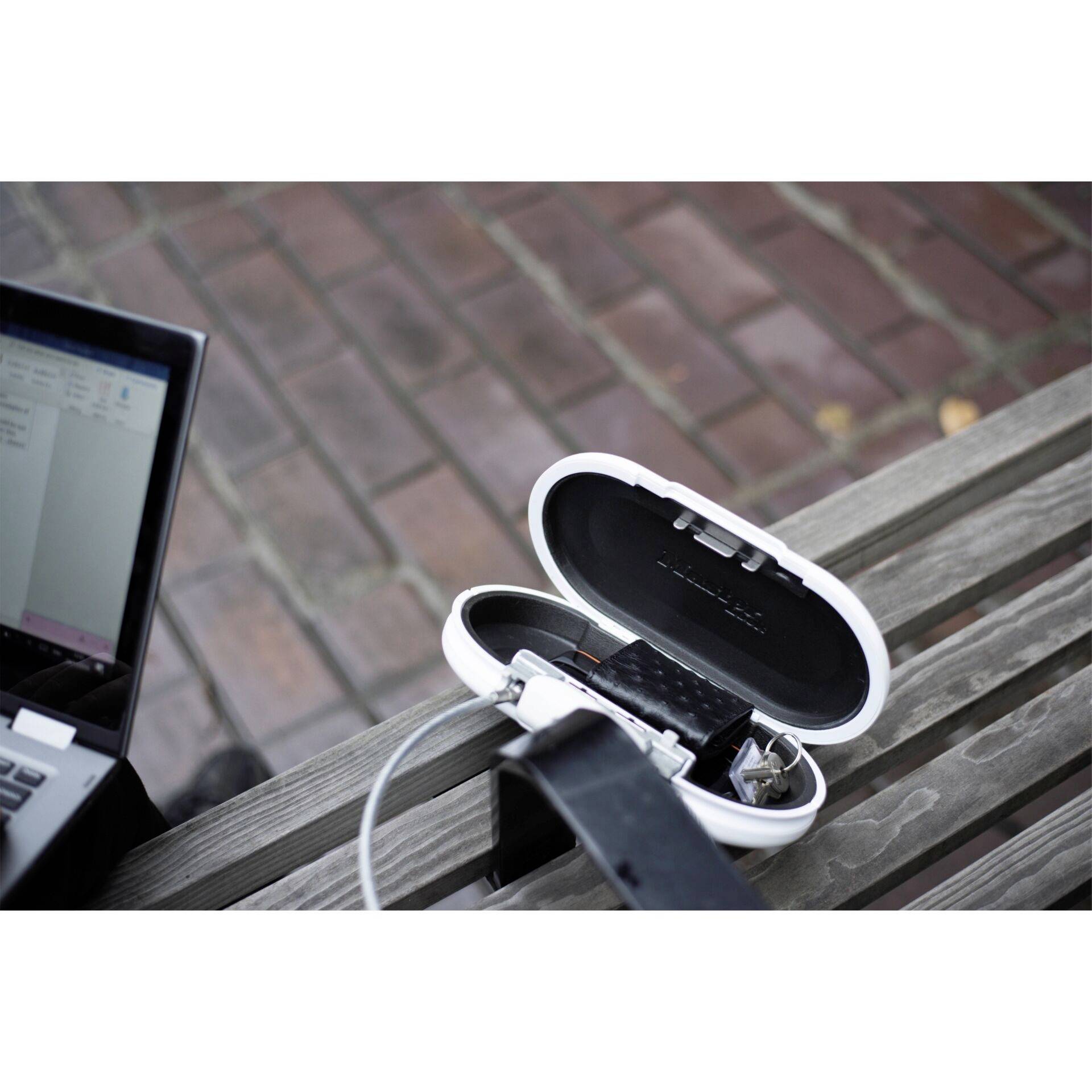 Ein Laptop lädt paar Kopfhörer in einer weißen Box auf einer Holzbank auf. Im Hintergrund sind Pflastersteine sichtbar.