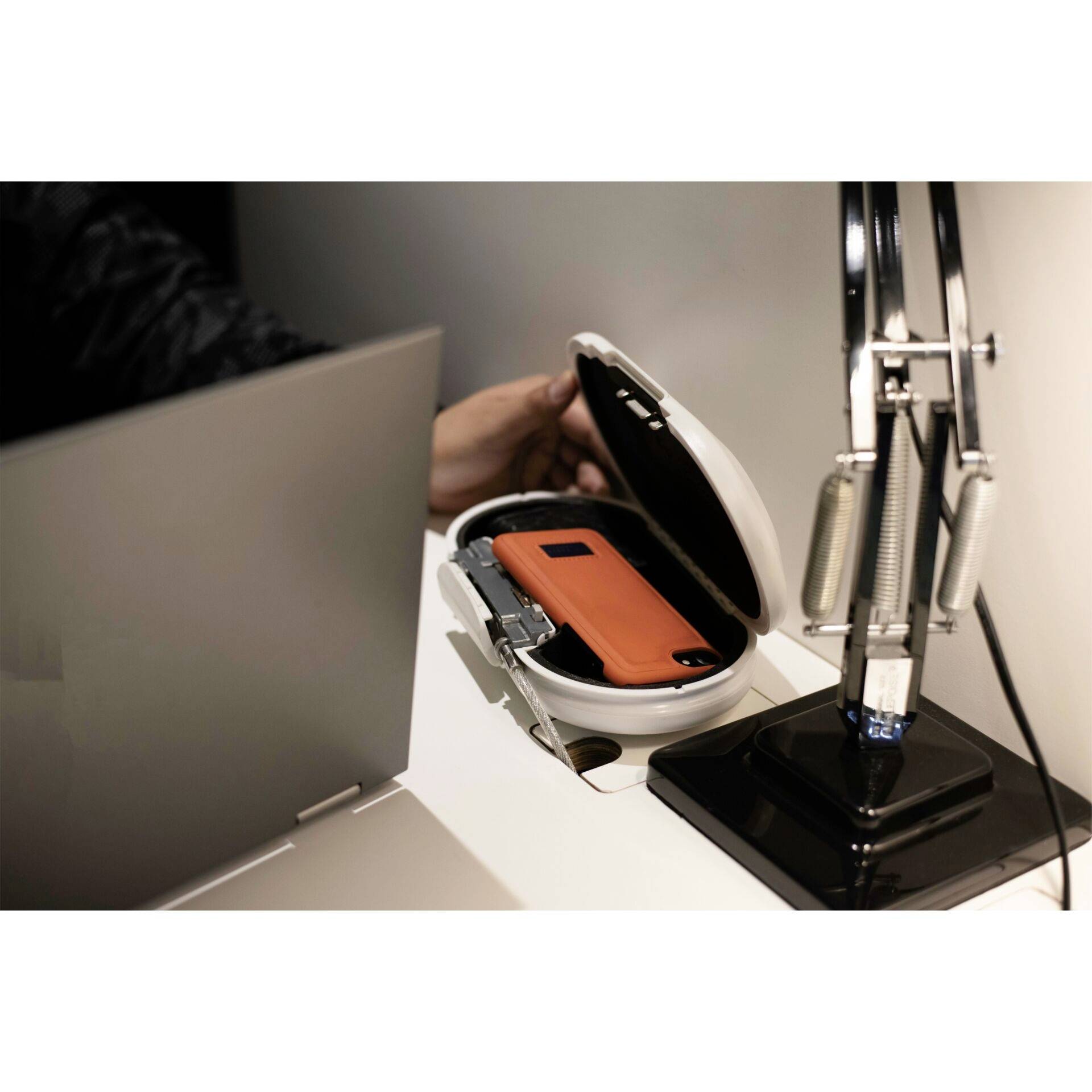 Eine Person desinfiziert ein Smartphone in einem runden UV-Gerät auf einem Tisch. Daneben stehen eine Schreibtischlampe und ein Laptop.