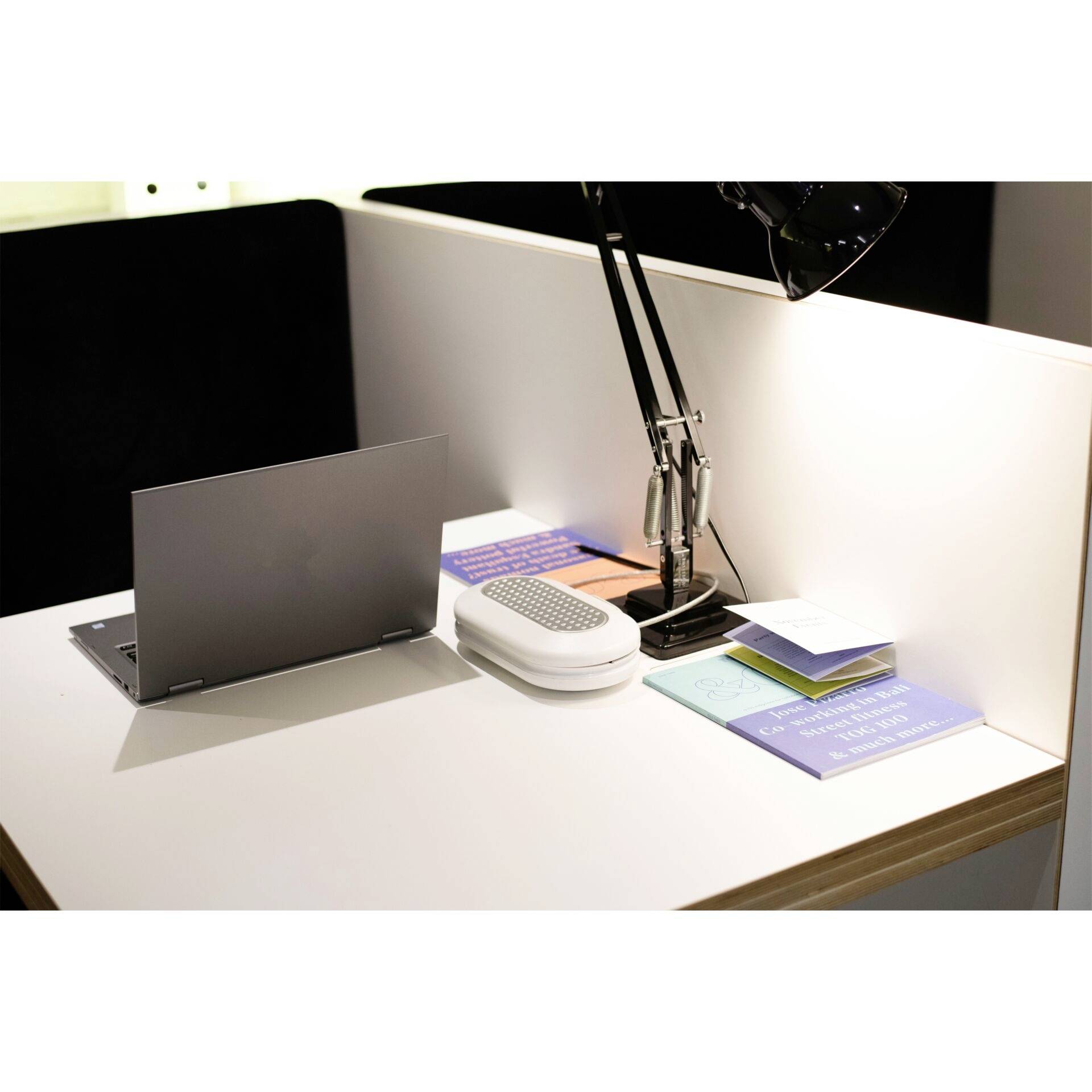 Arbeitsplatz mit aufgeklapptem Laptop, Lampe, Notizbüchern und Büchern auf einem Tisch. Die Umgebung ist modern und aufgeräumt.