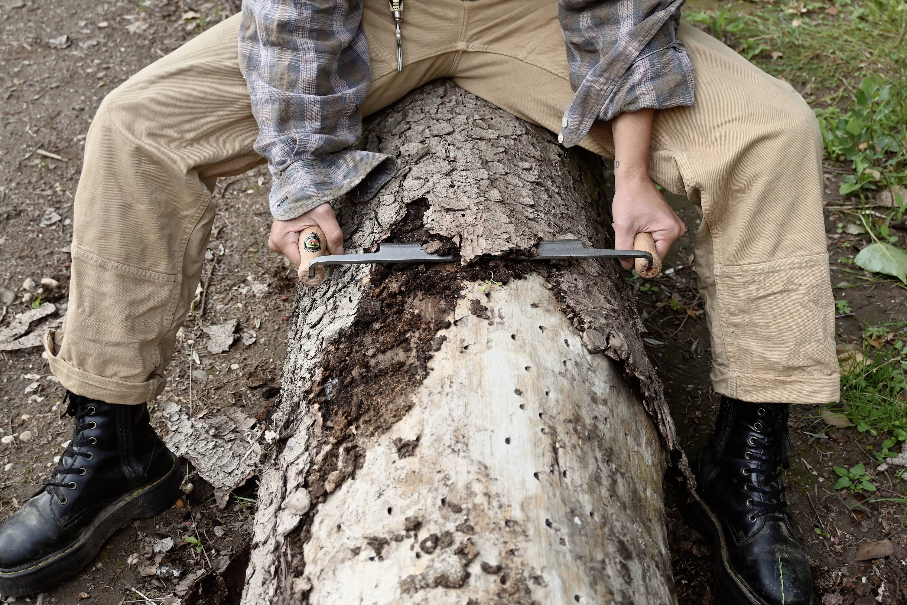Person sitzt auf einem Baumstamm und entfernt Rinde mit einem Zugmesser. Trägt Lederstiefel und kariertes Hemd.