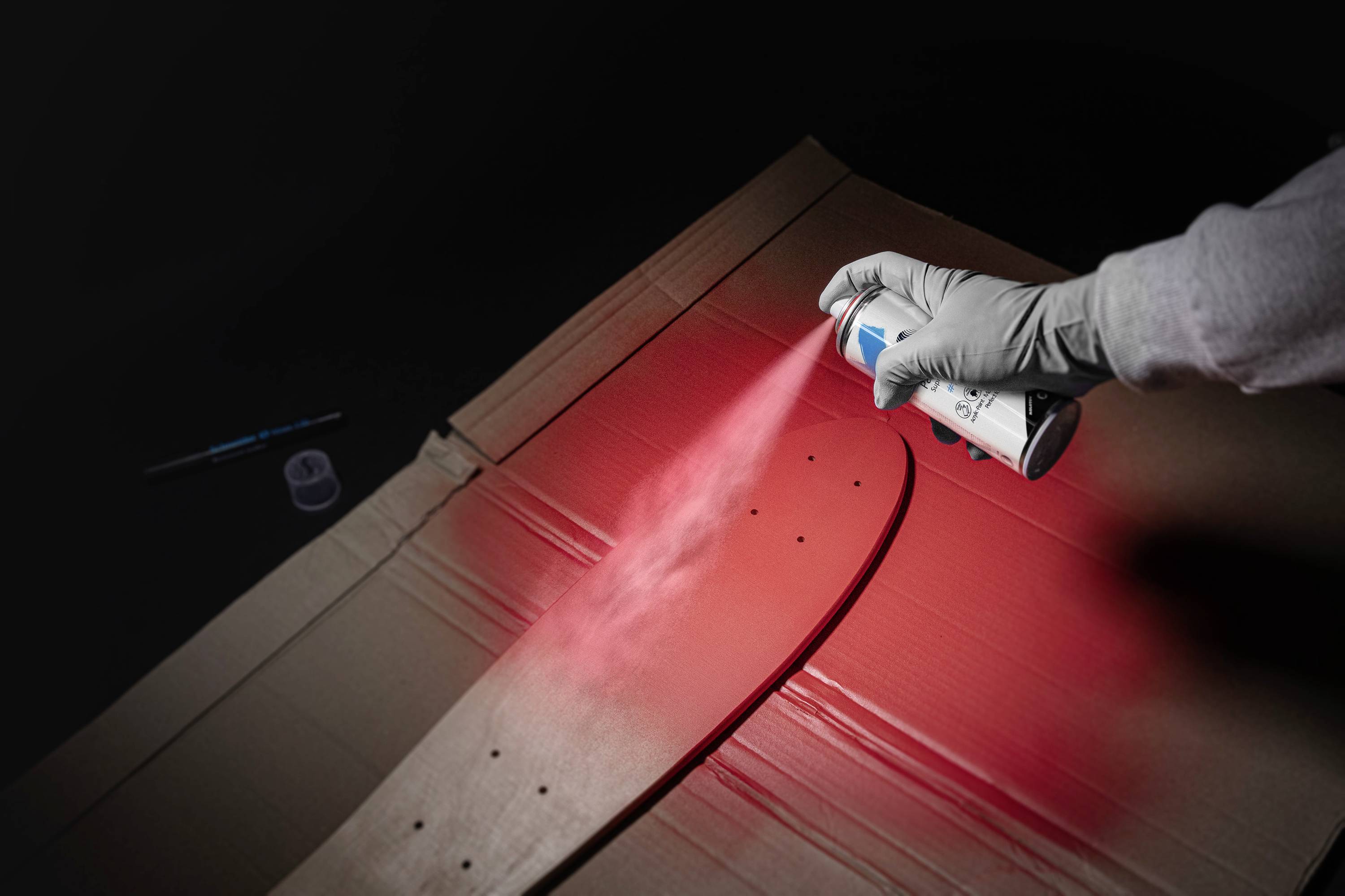 Eine Hand sprüht rote Farbe aus einer Sprühdose auf ein Skateboarddeck, das auf einem Karton liegt.