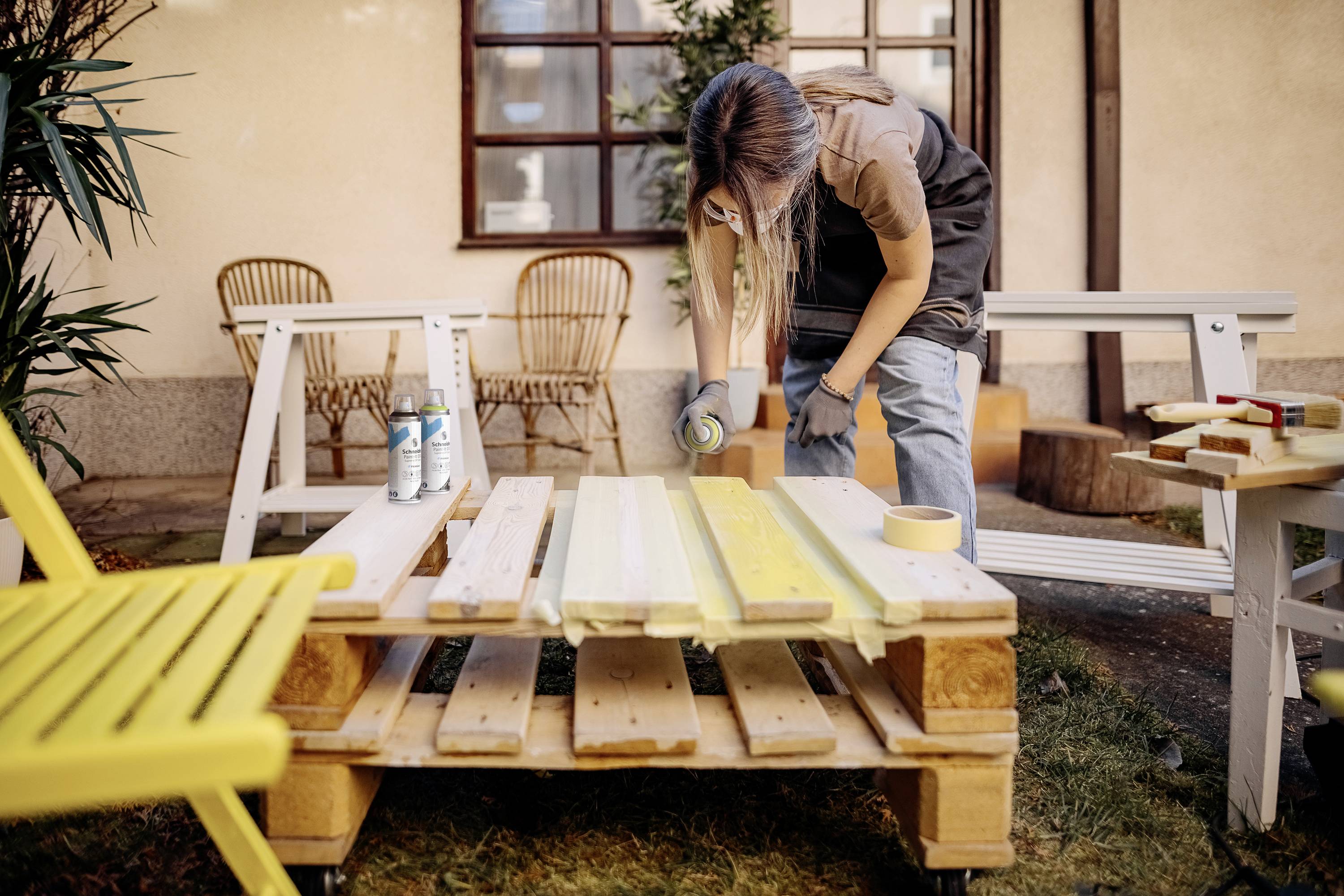Eine Person lackiert Holzbretter mit gelber Farbe auf einer Palette im Garten, umgeben von Pflanzen und Gartenmöbeln.