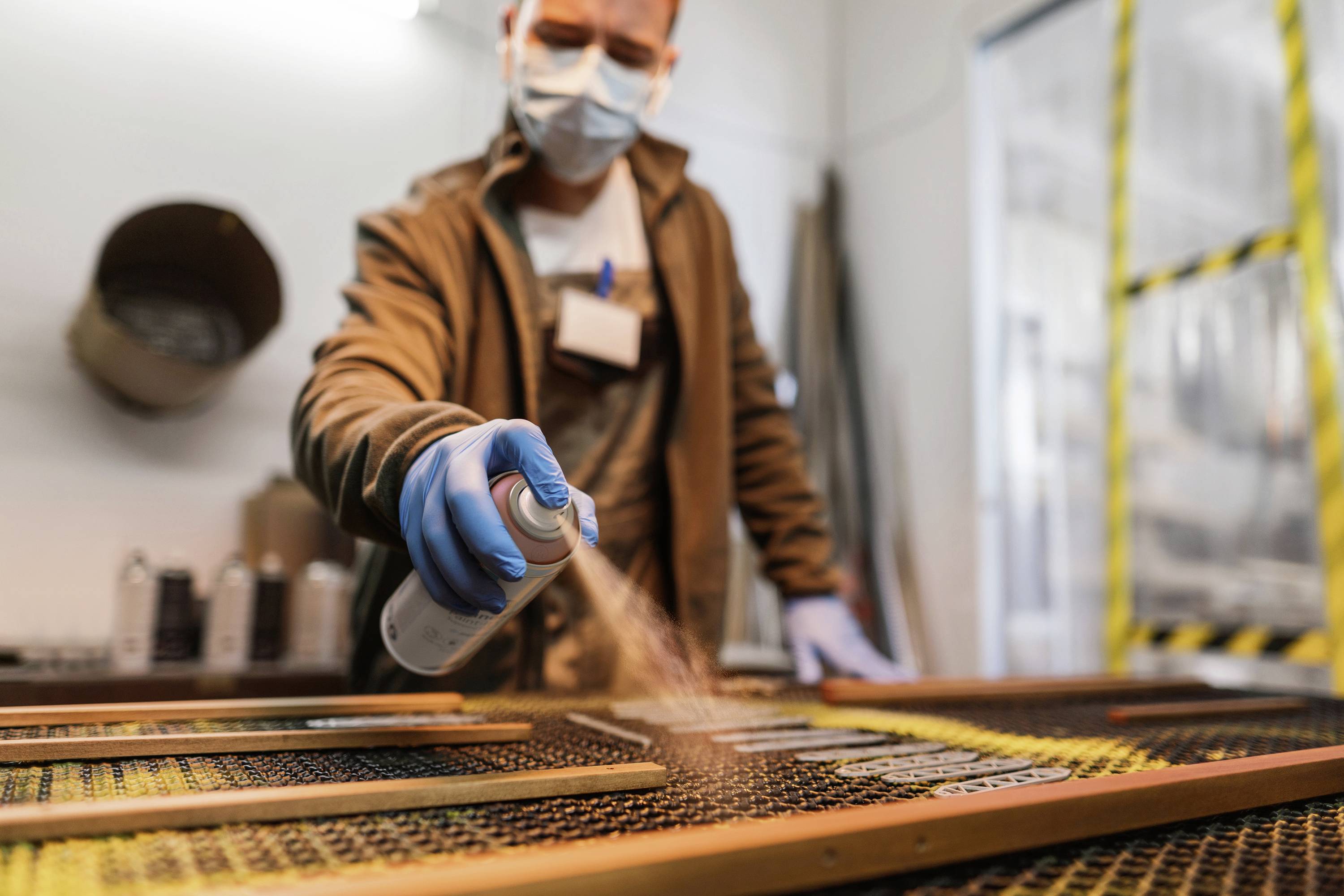 Eine Person mit Maske sprüht Farbe auf einen metallischen Gegenstand in einer Werkstatt. Im Hintergrund sind Werkzeuge sichtbar.