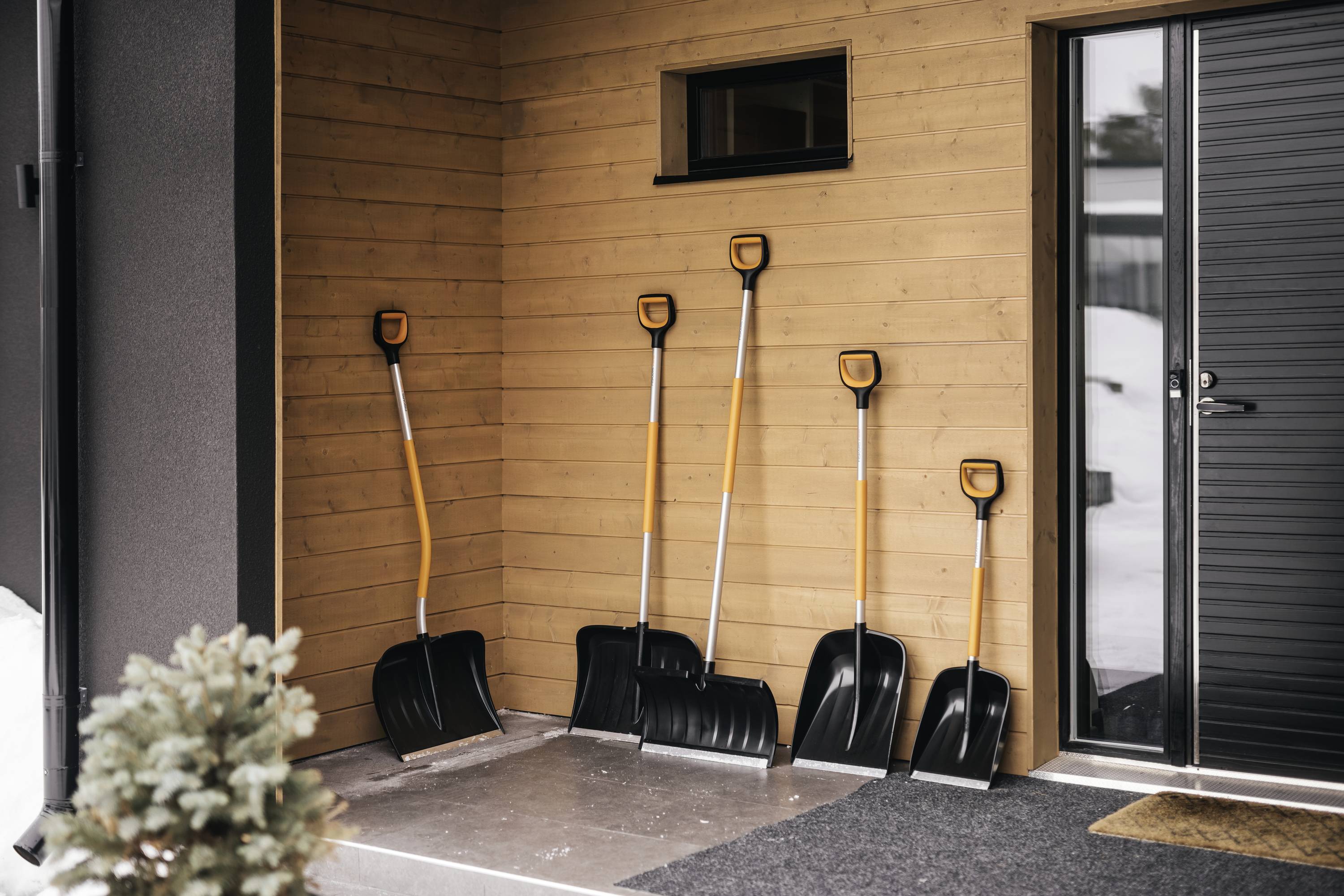 Fünf Schneeschaufeln lehnen an einer Holzwand neben einer Tür. Ein Baum ist im Vordergrund sichtbar, Schnee befindet sich draußen.