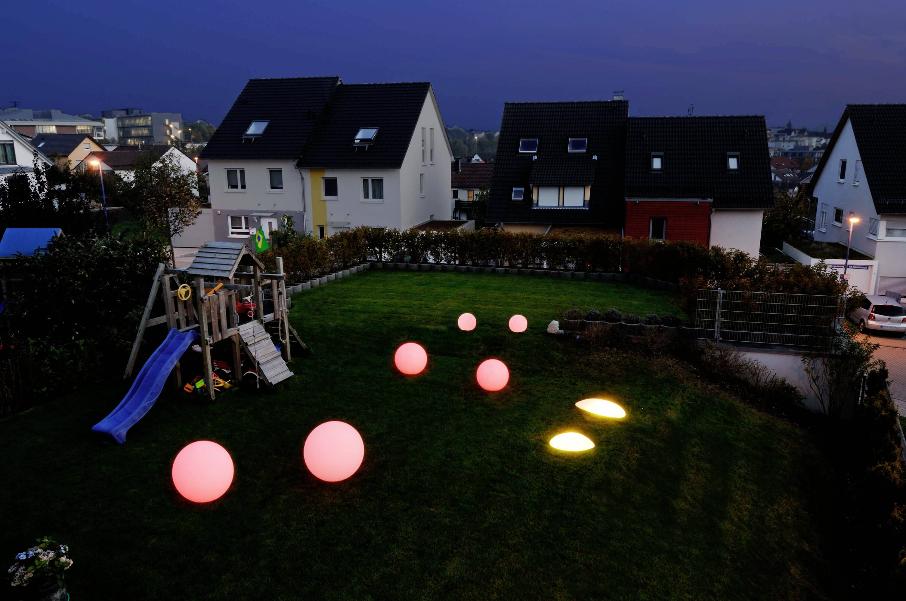 Ein Garten bei Dämmerung mit mehreren leuchtenden Kugeln auf dem Rasen. Im Hintergrund sind Häuser und ein Kinderspielplatz zu sehen.