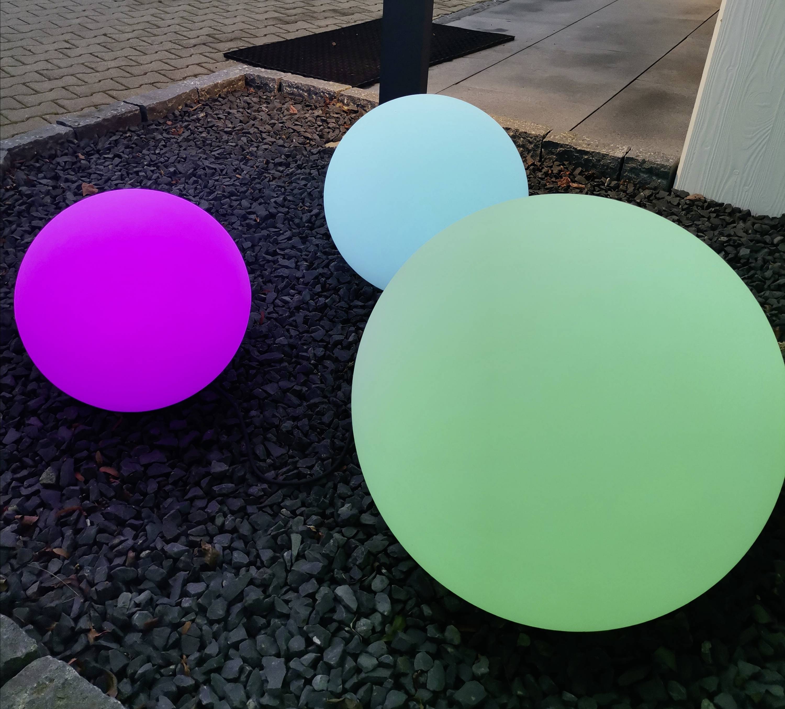 Drei bunte, leuchtende Kugeln in den Farben Lila, Blau und Grün liegen auf einem mit dunklen Steinen bedeckten Boden im Freien.