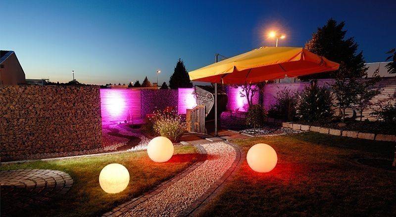 Ein Garten bei Nacht mit beleuchteten Kugeln auf dem Boden und einem großen gelben Sonnenschirm. Eine Steinmauer ist im Hintergrund.