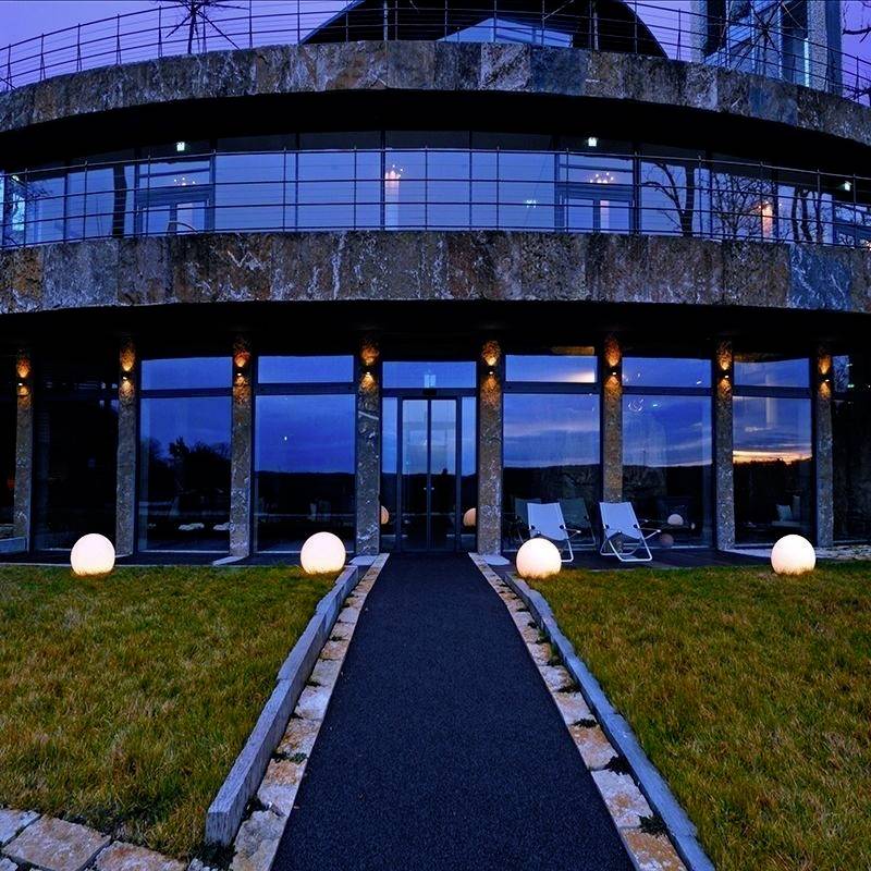 Ein modernes Gebäude mit großer Glasfassade und beleuchteten Kugelleuchten auf einem Rasen. Zwei Liegestühle stehen auf der Terrasse.