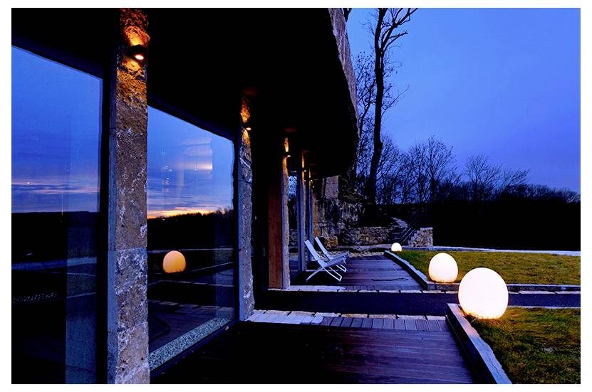 Moderne Terrasse neben einem Haus in der Abenddämmerung, mit leuchtenden kugelförmigen Lampen, Liegestühlen und großen Glasfenstern, die den Sonnenuntergang spiegeln.