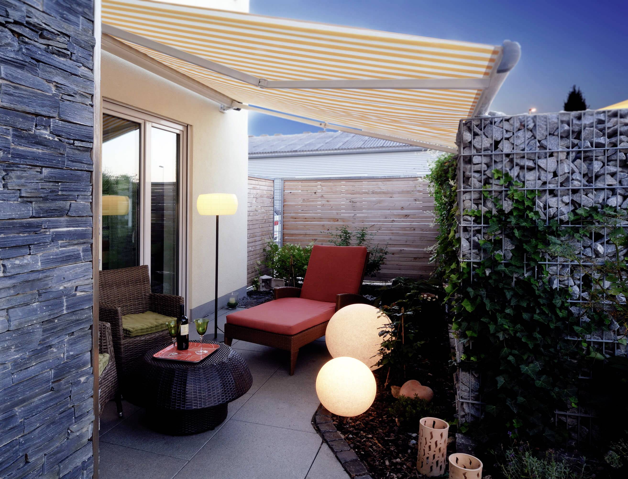 Eine gemütliche Terrasse mit Sitzgarnitur, Lampe und Pflanzendekor unter einer gestreiften Markise bei Abenddämmerung.