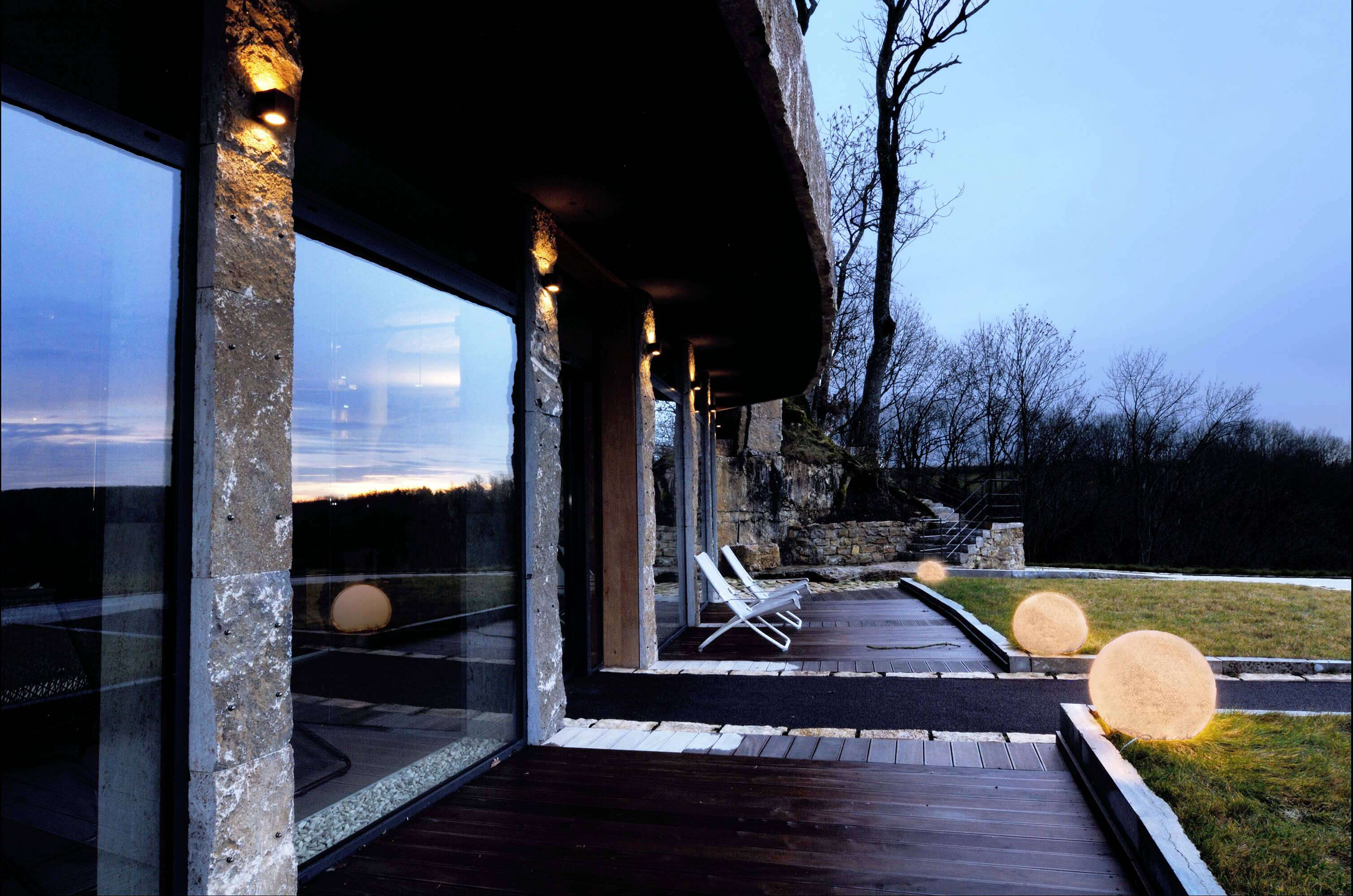 Ein modernes Gebäude mit großer Glasfassade und Holzterrasse bei Abenddämmerung. Im Hintergrund Bäume, im Vordergrund leuchtende Kugeln.